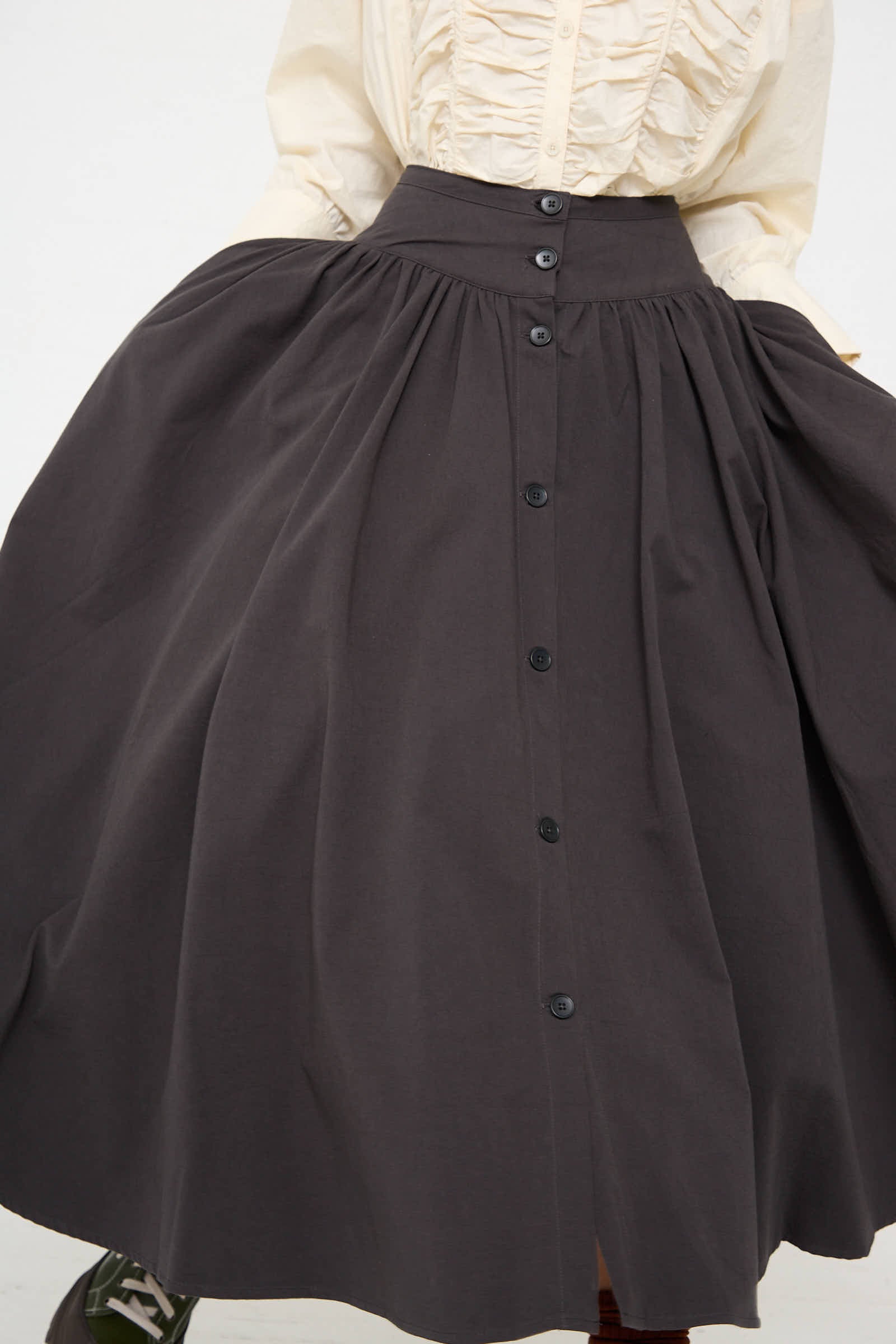 A person is wearing the Black Crane Woven Cotton Circular Skirt in Dark Grey, which has a high-waisted design, black buttons down the front, a wide-banded waist, and an A-line silhouette, paired with a light-colored ruffled blouse.