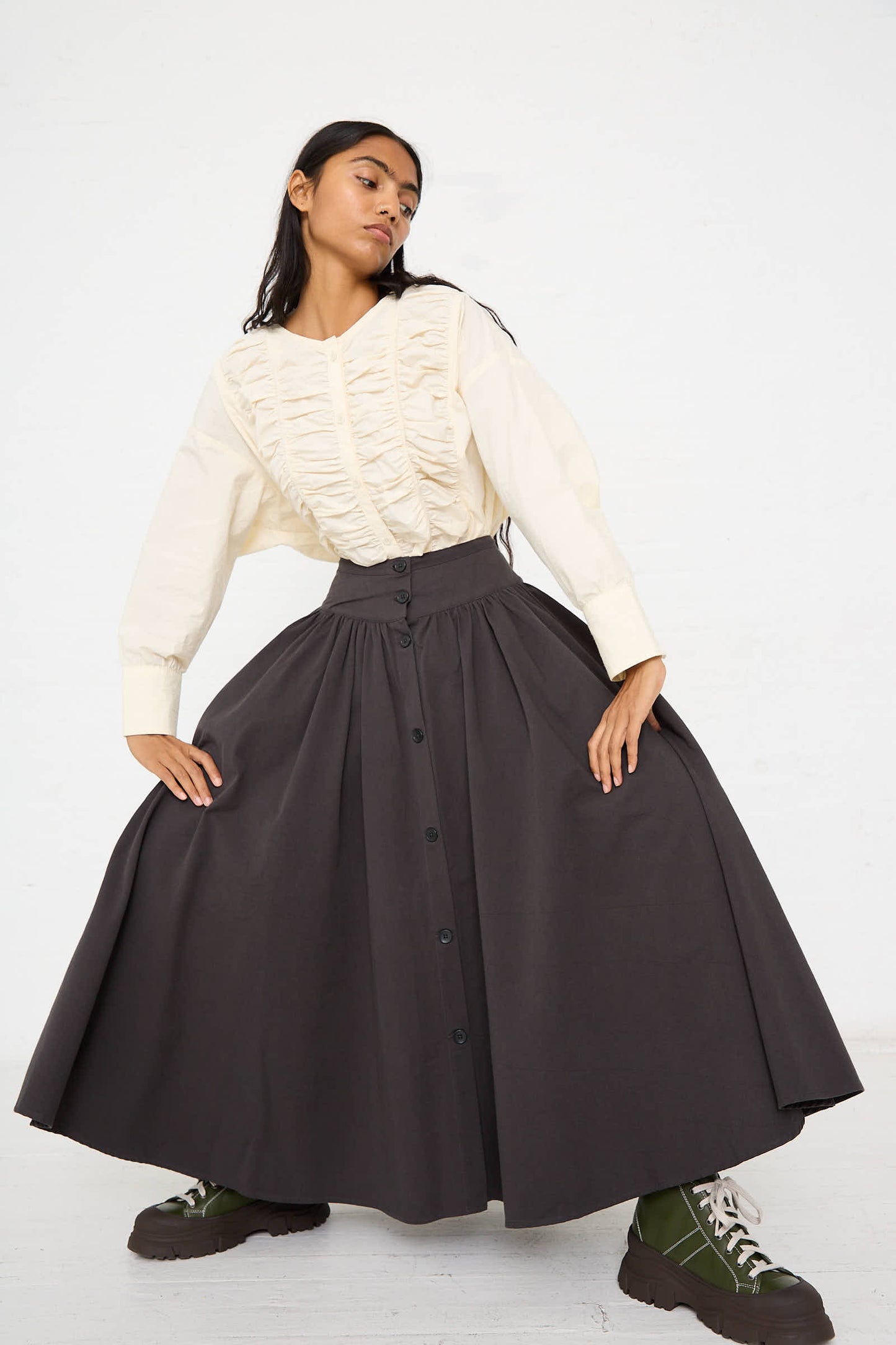 A person stands with a wide stance, wearing an off-white ruffled blouse tucked into Black Crane's Woven Cotton Circular Skirt in Dark Grey, which features a wide banded waist and an elegant a-line silhouette, paired with green boots against a plain white background.