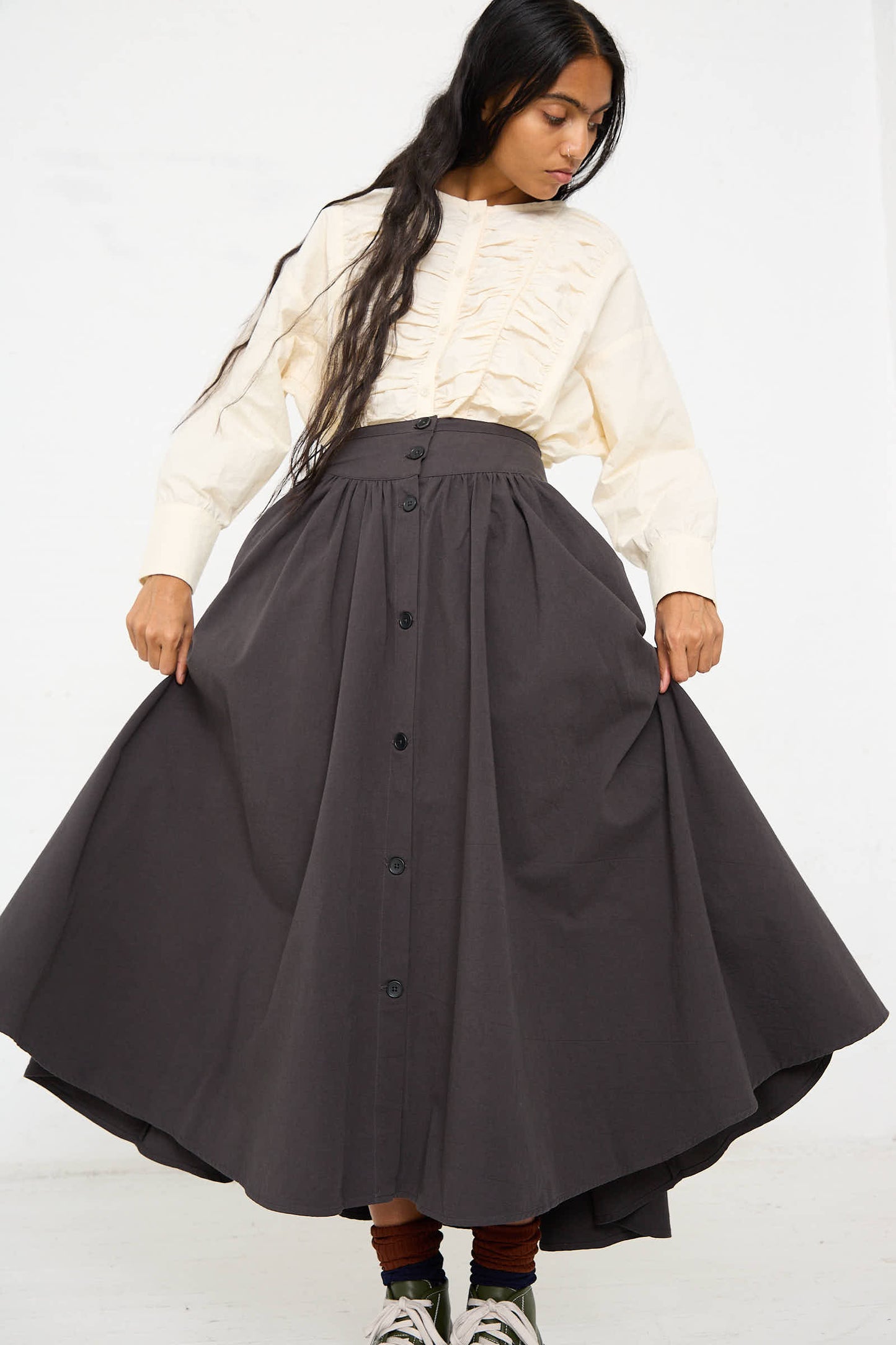 A person is wearing a cream-colored blouse with ruffled details and a Woven Cotton Circular Skirt in Dark Grey from Black Crane, featuring a wide banded waist and button-front design. They are holding out the sides of the skirt against a plain white background.