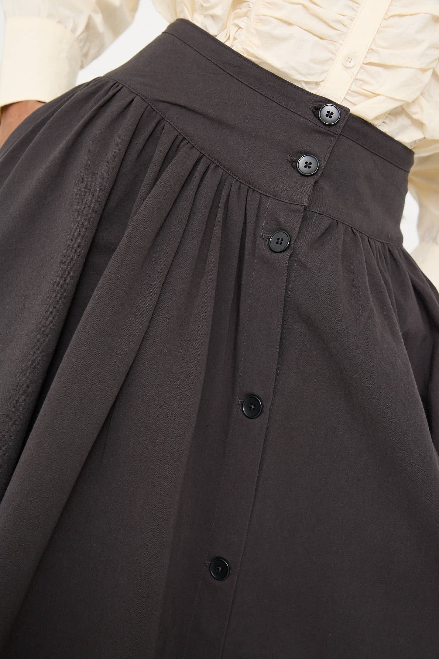 A close-up of a person wearing a beige, long-sleeved shirt tucked into a high-waisted Black Crane Woven Cotton Circular Skirt in Dark Grey, featuring an A-line silhouette with pleats and four buttons down the front. The skirt boasts a wide banded waist in woven Japanese cotton for added elegance.