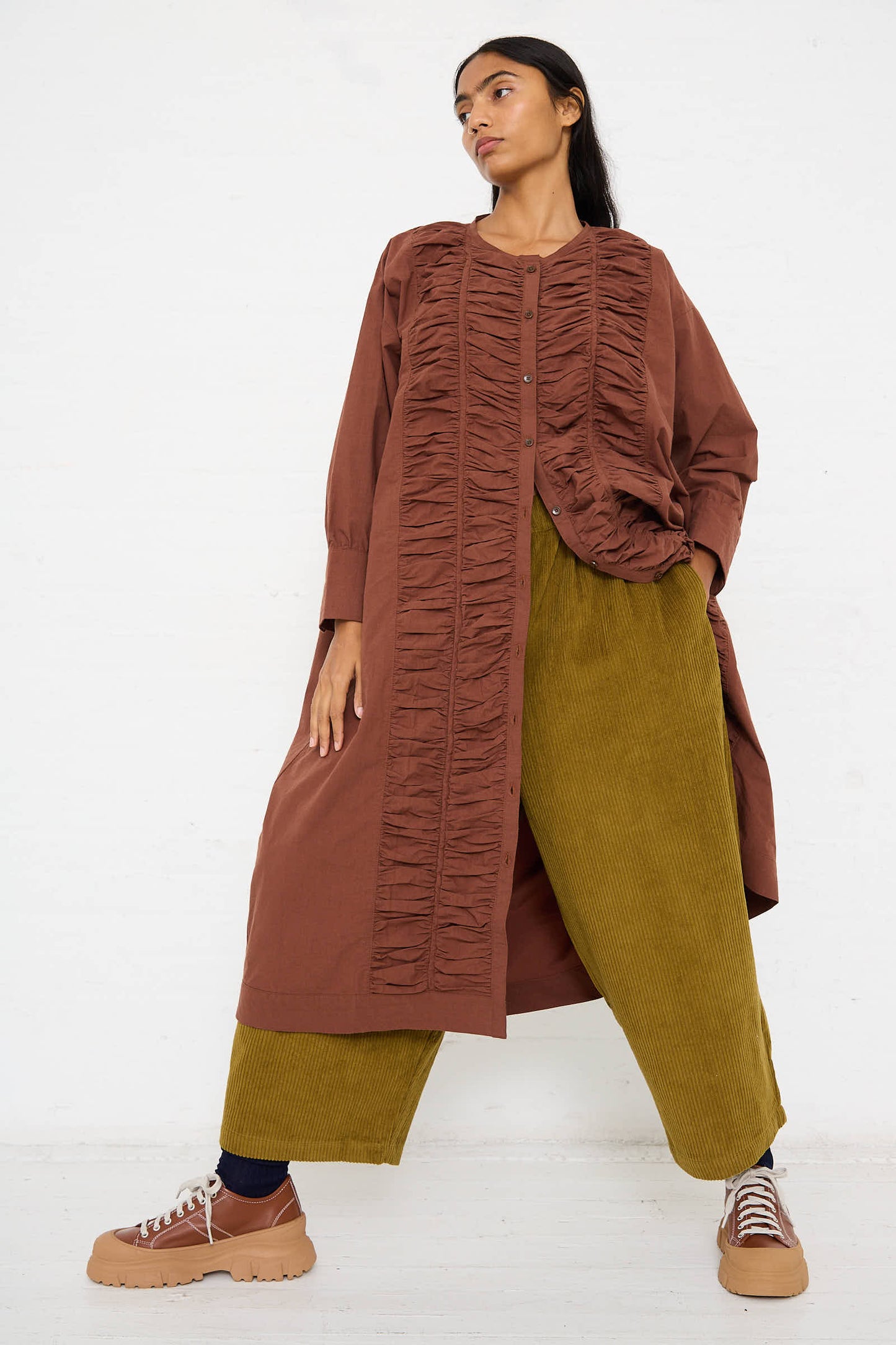 Someone with long hair wears a Woven Cotton Ruffled Dress in Cacao by Black Crane, along with green corduroy pants in a relaxed fit and beige sneakers, posing against a plain white background.