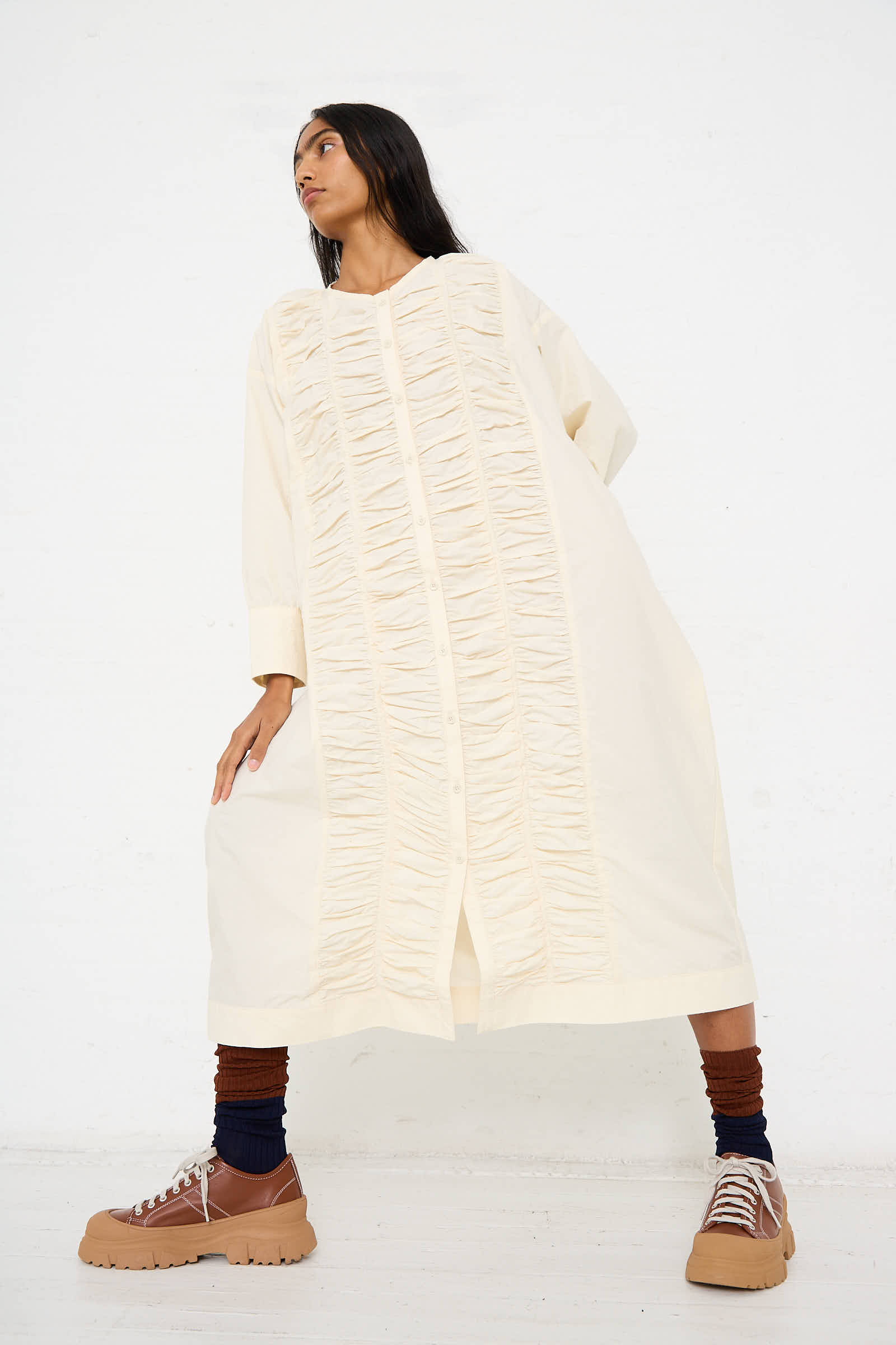 A woman in a relaxed fit, floor-length Woven Cotton Ruffled Dress in Ivory by Black Crane stands against a plain white background, looking to the side. She wears black and rust ankle socks and tan hiking boots.