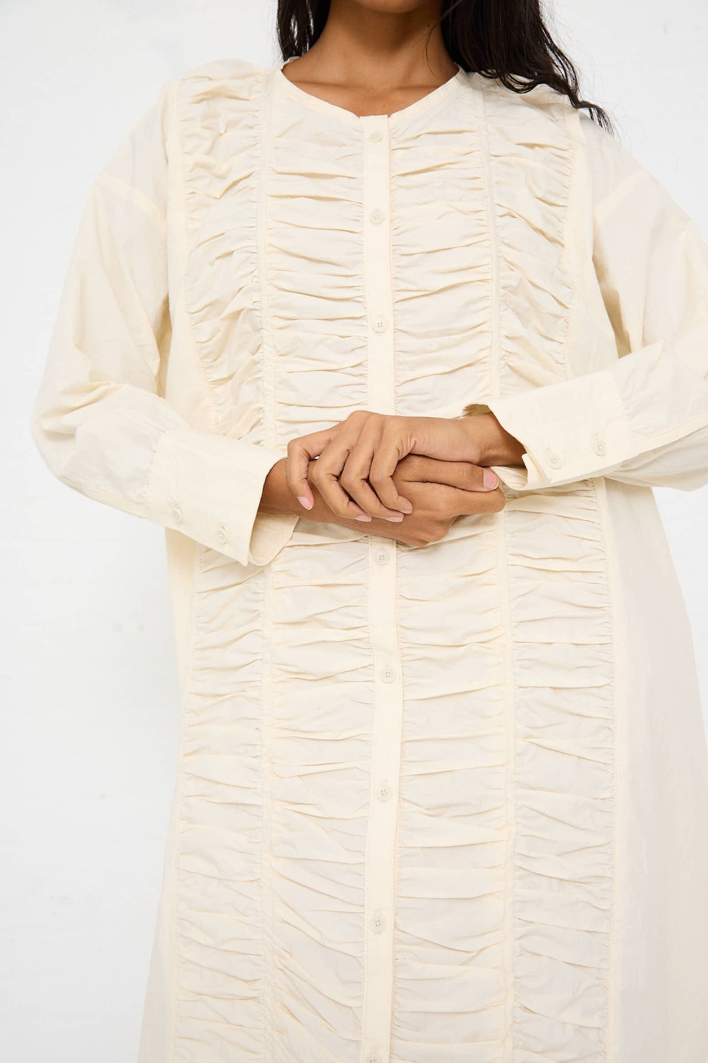 A person wearing the Black Crane Woven Cotton Ruffled Dress in Ivory with hands clasped in front.