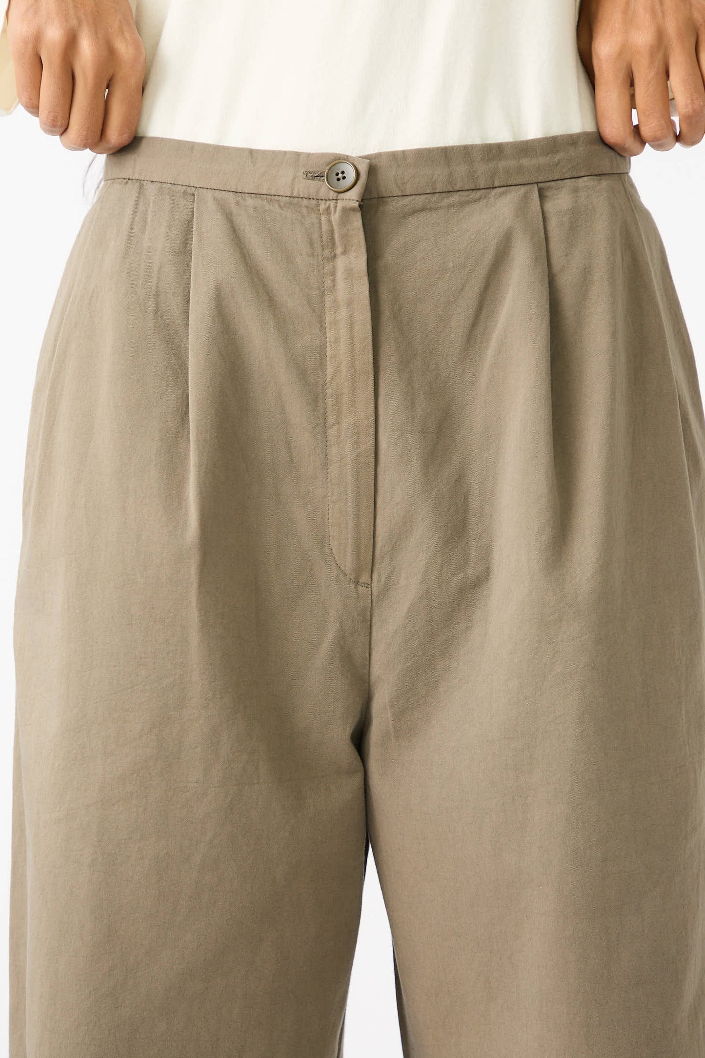 Close-up of a person wearing Black Crane's dark olive, Woven Cotton Straight Trousers with a button and fly closure. Crafted from Japanese cotton, these trousers feature a straight leg. The person is tucking in a white shirt, visible at the top of the image.