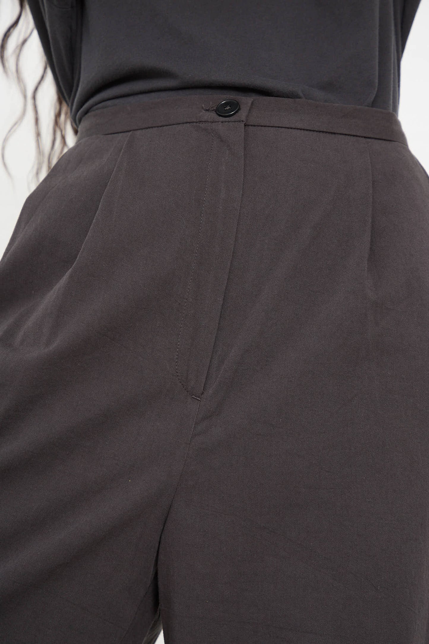 Close-up of a person wearing the Woven Cotton Straight Trouser in Dark Grey by Black Crane, crafted from woven Japanese cotton, featuring a button closure, pleats on the front, and a sleek tapered leg.