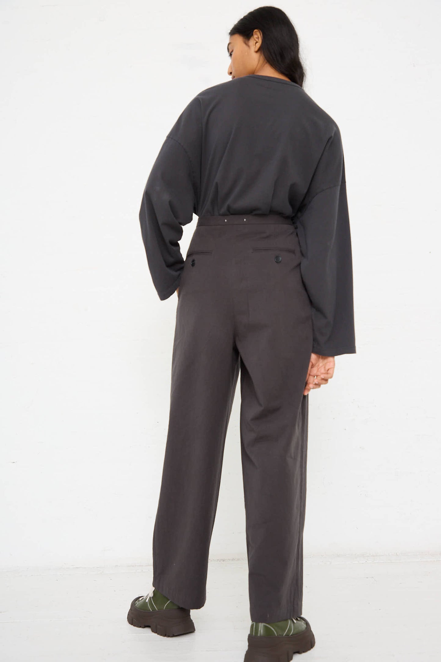 A person stands against a white background wearing a dark, oversized long-sleeve shirt and the Woven Cotton Straight Trouser in Dark Grey from Black Crane. They are turned away from the camera.