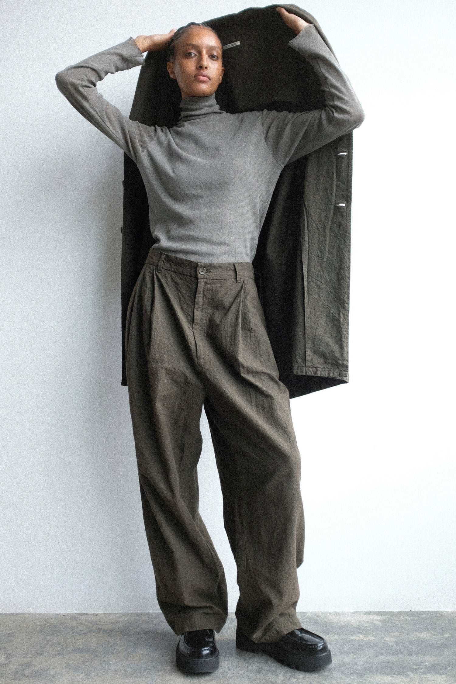 Person wearing a Black Crane's Crinkled Coat in Charcoal, Double Mesh Turtleneck in Dark Stone, and Crinkled Trousers in Charcoal, against a white background.