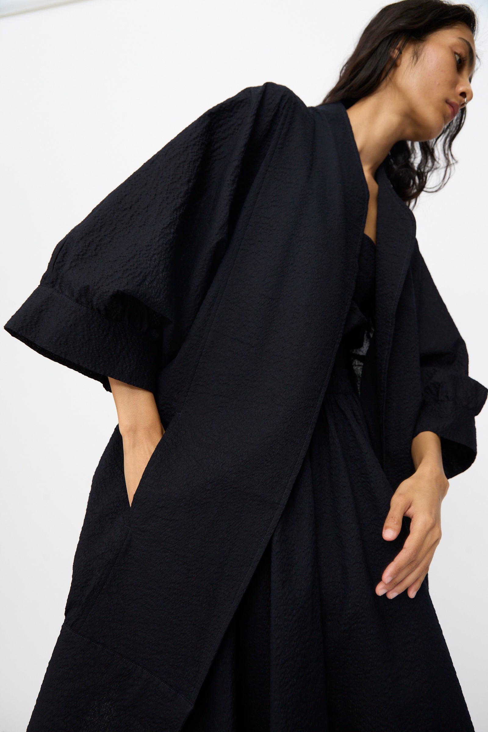 A woman wears the Black Crane Textured Cotton Spoon Jacket in Midnight—a long, oversized jacket with wide sleeves, side pockets, and a textured black Japanese cotton finish—as she stands against a plain white background.