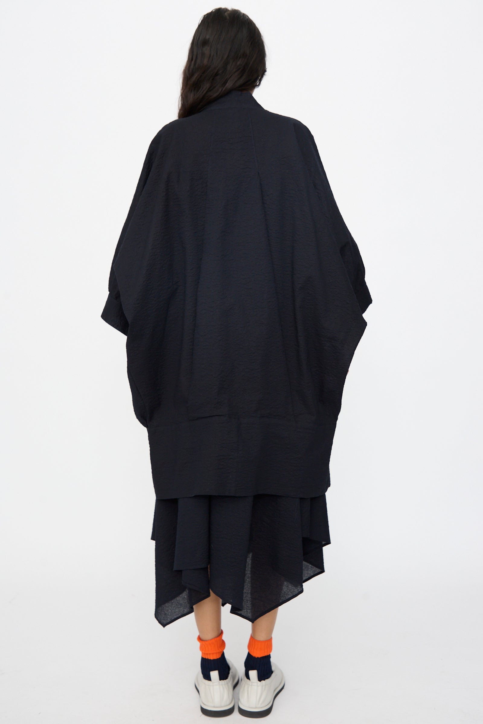 A person with long dark hair faces away, wearing the Black Crane Textured Cotton Spoon Jacket in Midnight, paired with white sneakers and orange and navy socks, set against a plain white background.