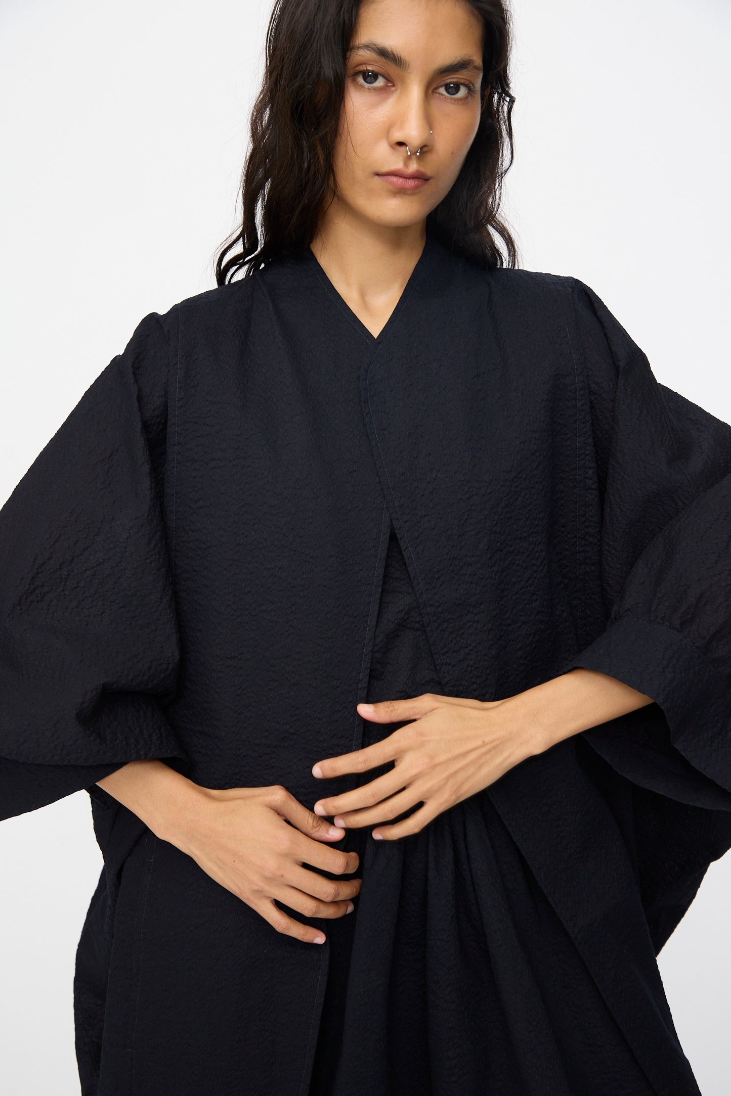 A person with long dark hair wears the Black Crane Textured Cotton Spoon Jacket in Midnight, styled kimono-like, and poses with hands over their abdomen against a plain background.