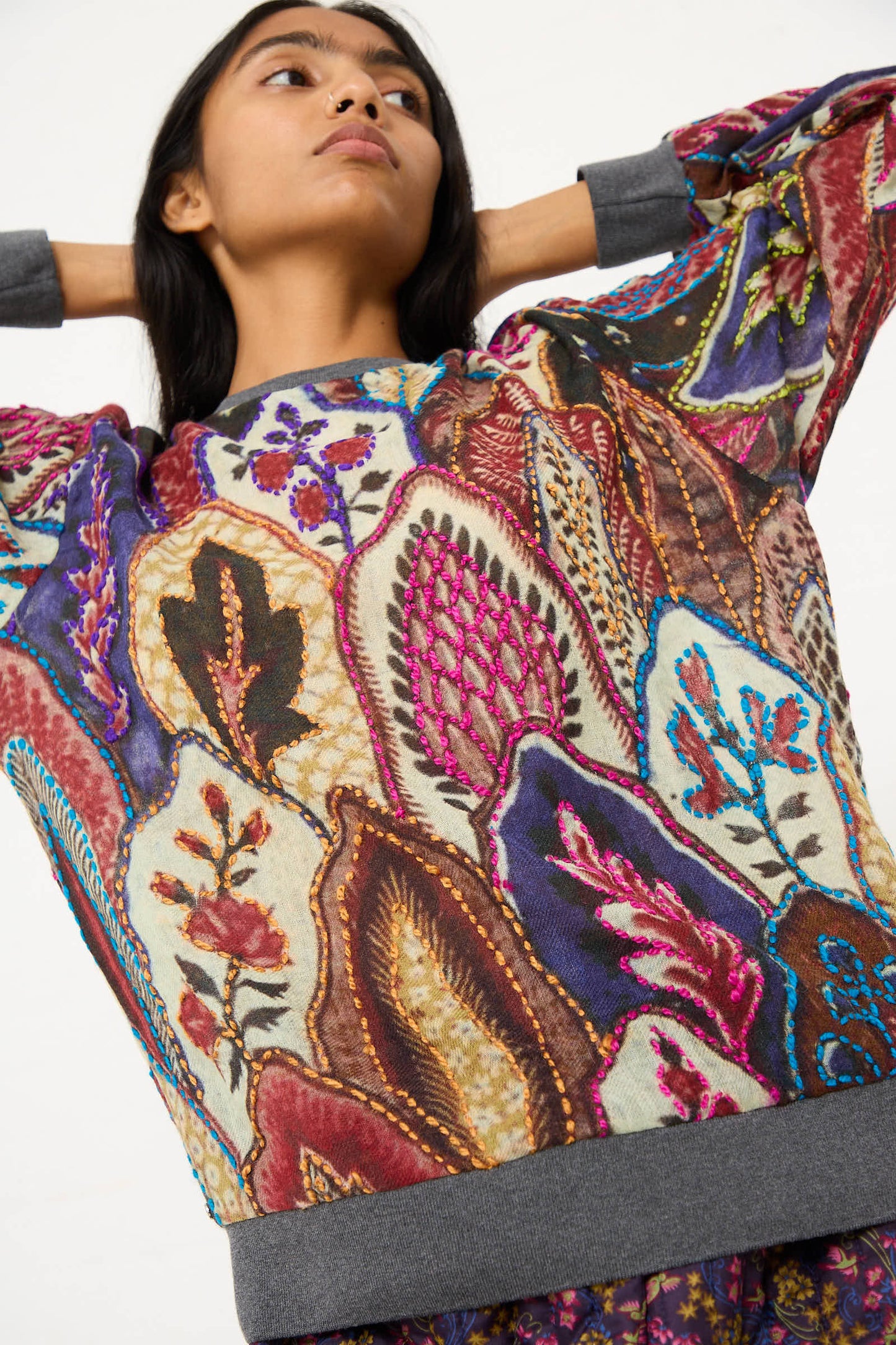 Person standing, arms raised, wearing the Bless Embroidered Wool No. 78 Sweater in Multicolor Brown with intricate patterns and various shapes in shades of purple, red, and brown.