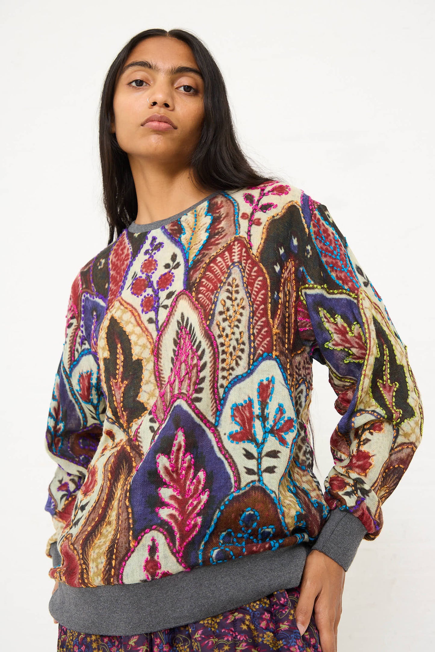 A person with long dark hair wears the Bless Embroidered Wool No. 78 Sweater in Multicolor Brown, featuring floral motif embroidery, paired with patterned pants. The background is plain white.