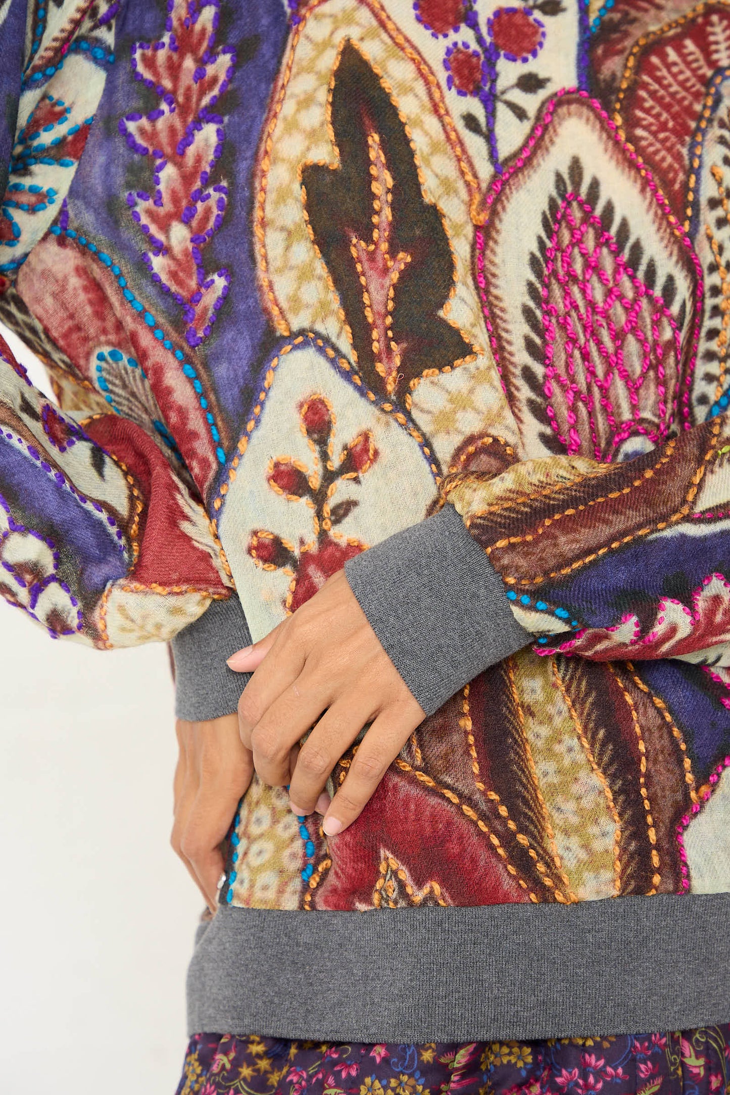 A person wearing the Bless Embroidered Wool No. 78 Sweater in Multicolor Brown, with a colorful and intricate leaf and floral pattern, their hands lightly clasped in front of them.