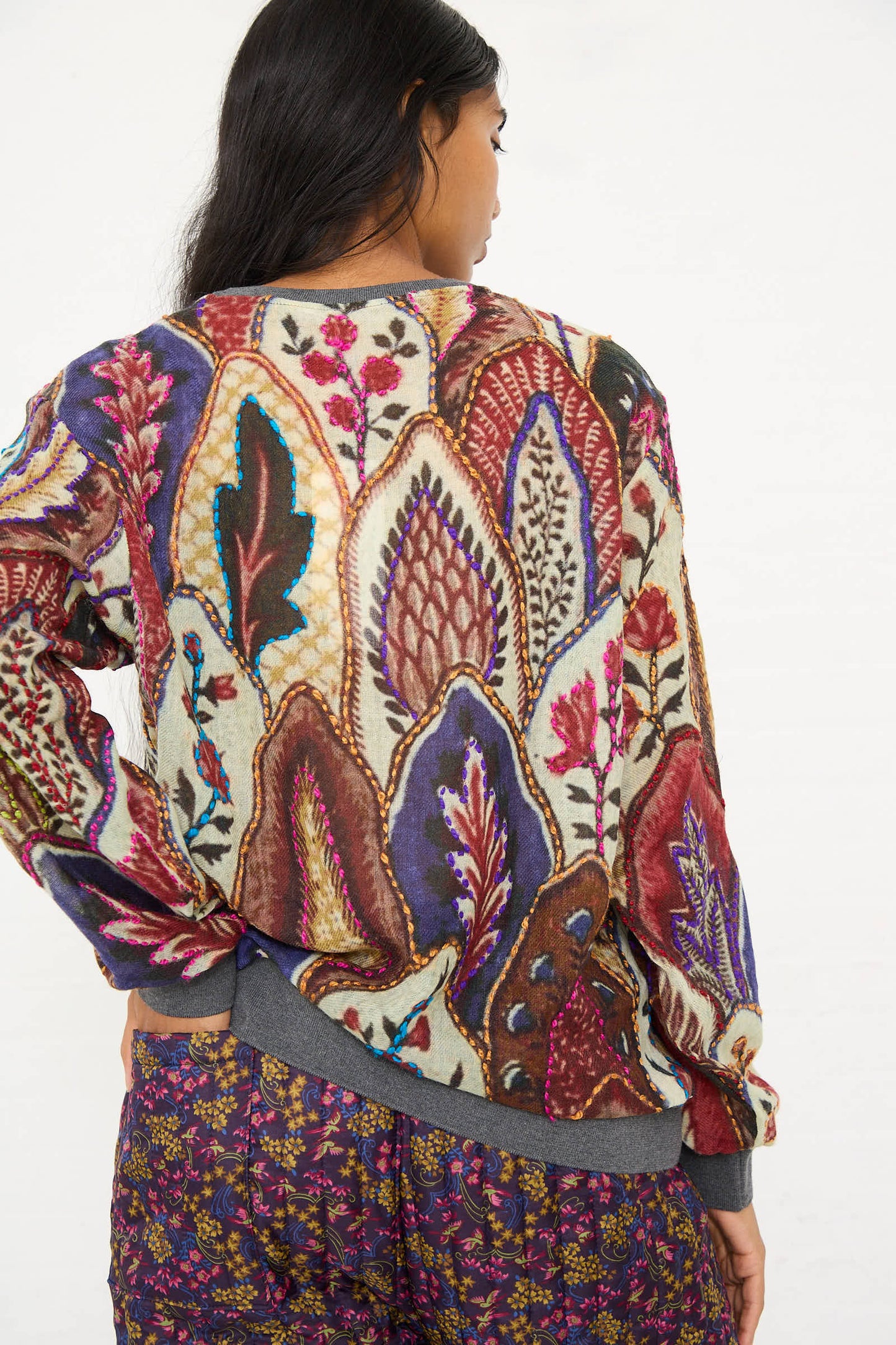 A person with long hair wears the Bless Embroidered Wool No. 78 Sweater in Multicolor Brown, featuring intricate floral motif embroidery and leaf designs. They are paired with relaxed fit patterned pants. The person is turned away, showcasing the detailed back of the sweater.