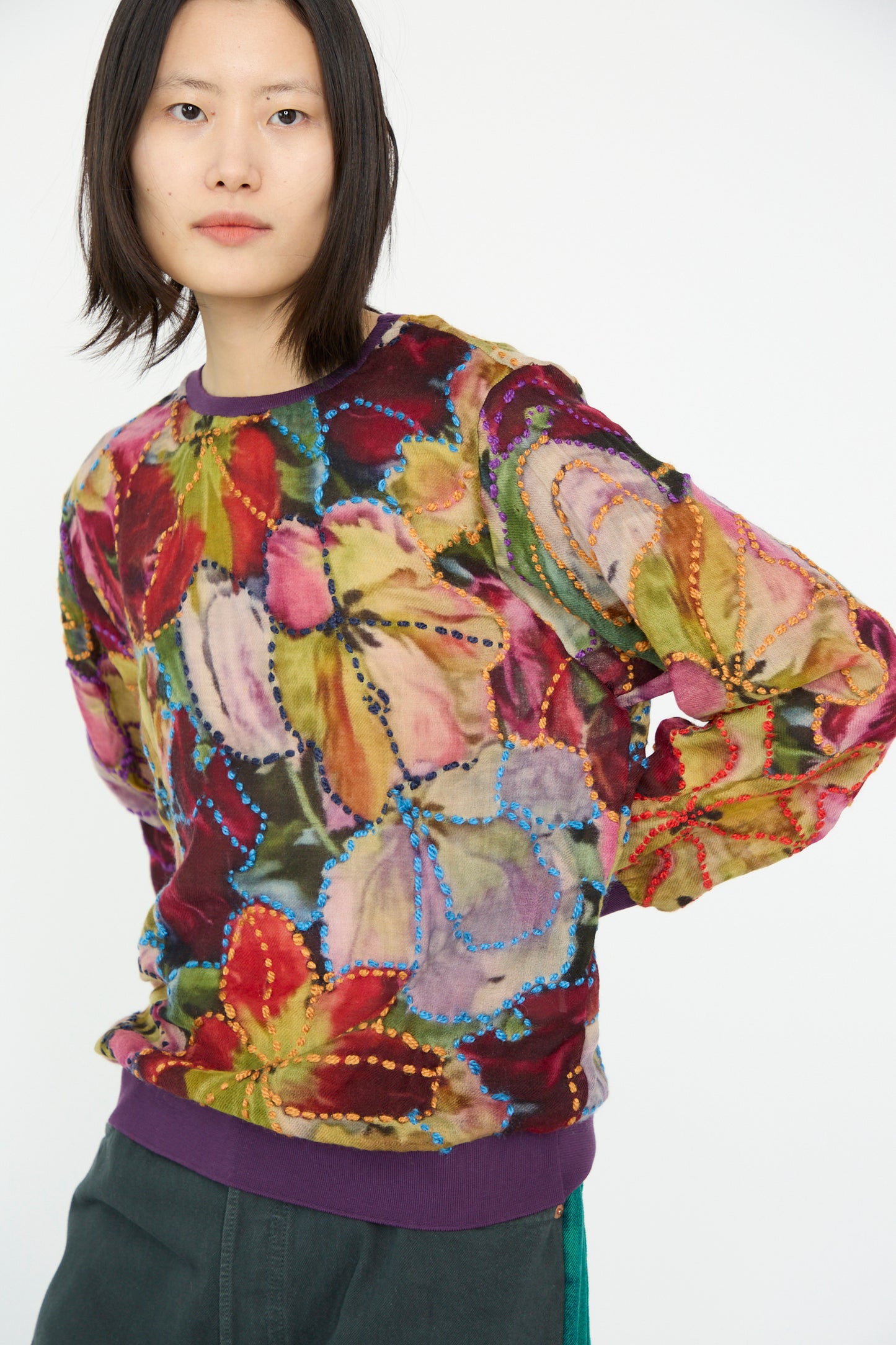 A person wearing the Embroidered Woolmix No. 78 M-Sweater in Multi by Bless, featuring a relaxed fit with colorful floral patterns and vivid purple cuffs, pairs it with dark green pants against a plain background.