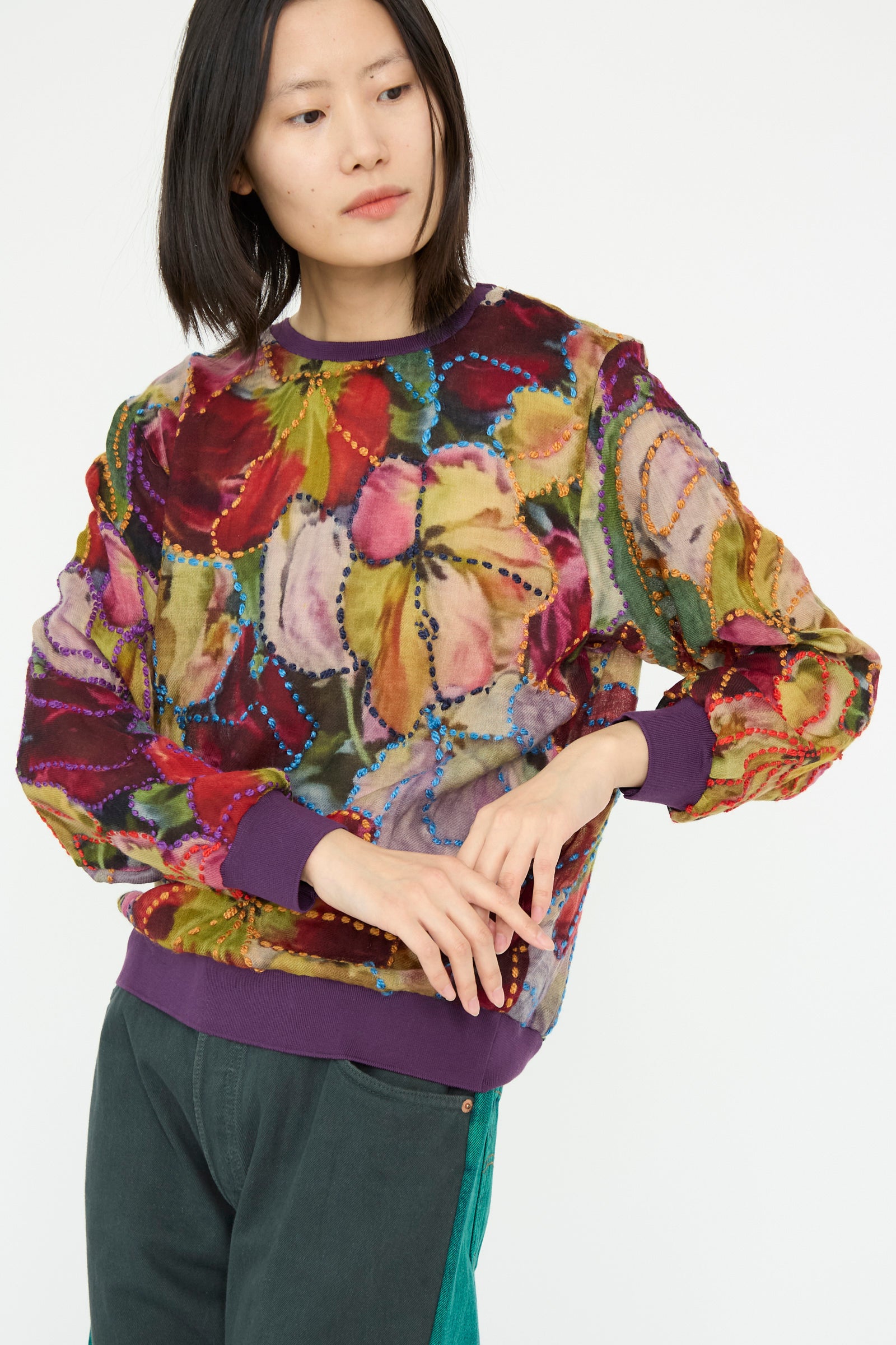 A model stands against a plain background wearing the Bless Embroidered Woolmix No. 78 M-Sweater in Multi paired with dark pants and hands clasped at the front.