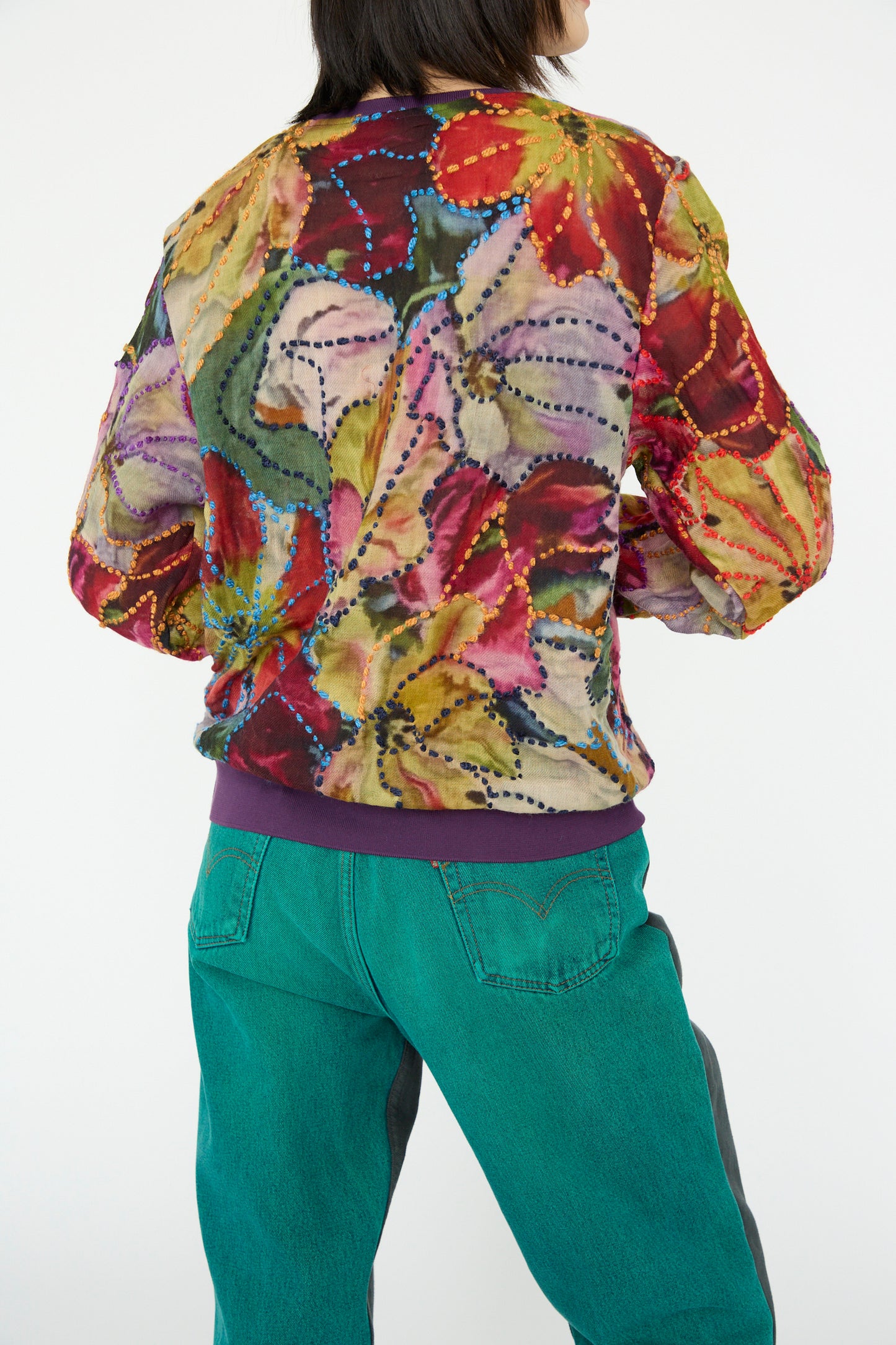 A model in a Bless Embroidered Woolmix No. 78 M-Sweater in Multi and teal pants stands facing away, highlighting vibrant colors.