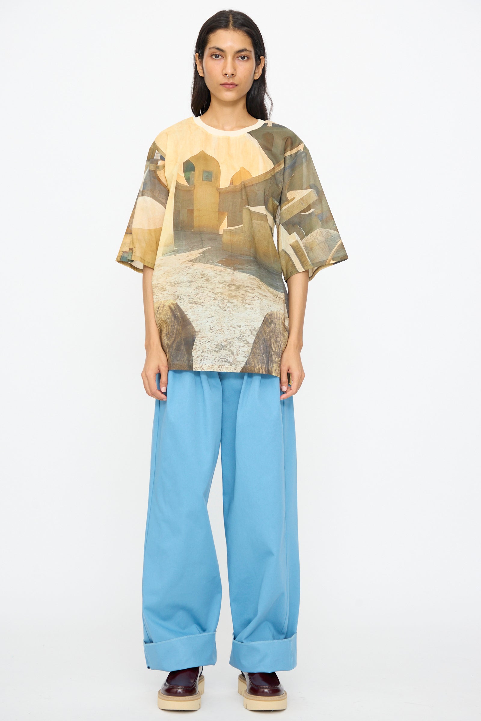 A person stands against a white background wearing the Bless Holidayjaipurobservatory Tee in Print, featuring an oversized boxy fit and landscape design, paired with loose light blue pants and dark platform shoes.