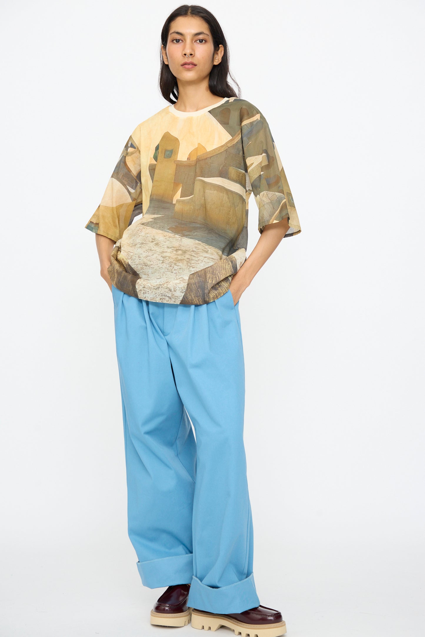 A person stands against a white background, wearing the Bless Holidayjaipurobservatory Tee in Print—a semi-sheer, oversized boxy cotton top with an abstract design—paired with loose light blue pants and maroon platform shoes.