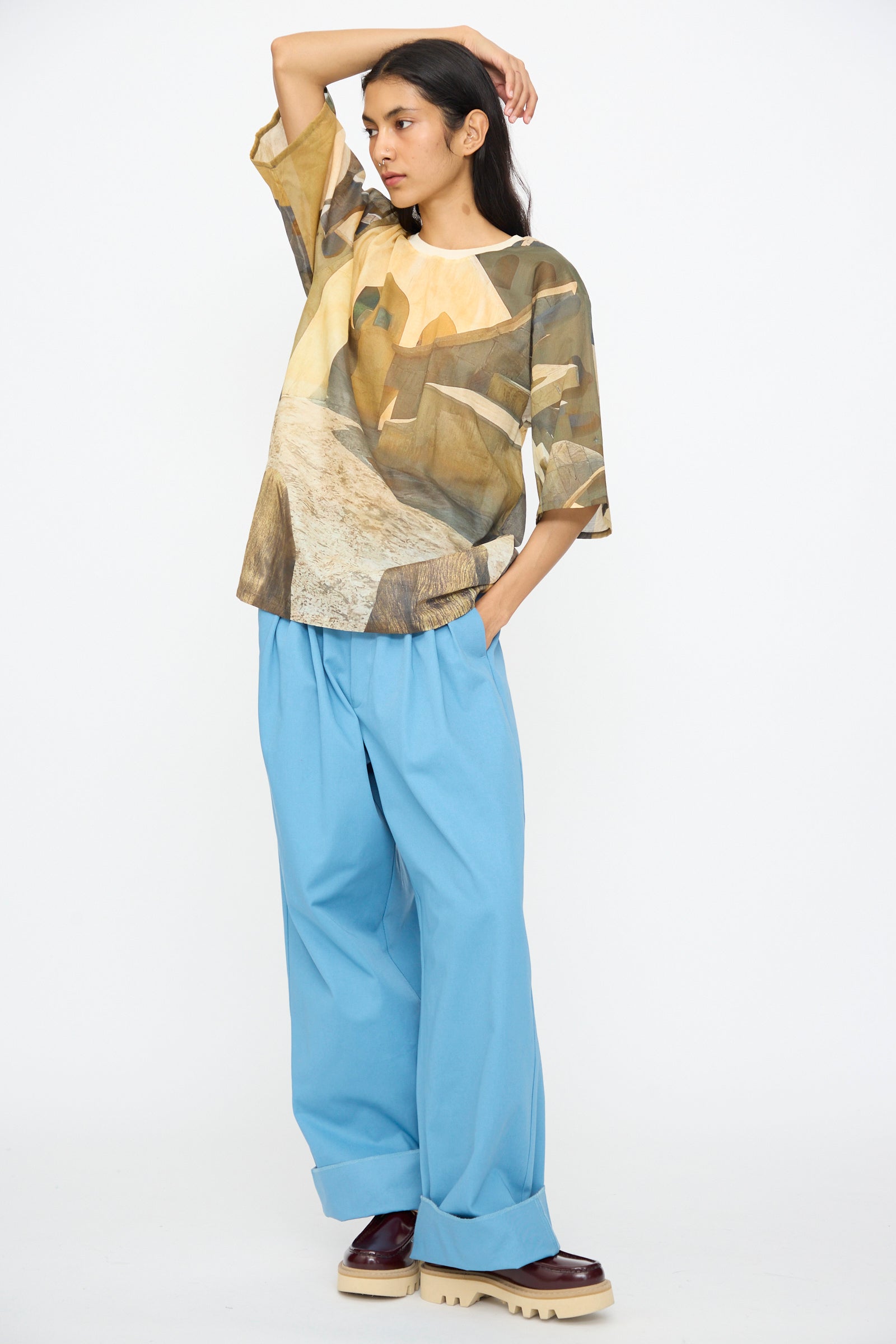 Wearing Bless’s Holidayjaipurobservatory Tee in Print, a person stands against a white background in an oversized boxy top, wide light blue trousers, and chunky shoes, one hand in pocket and the other touching their head.