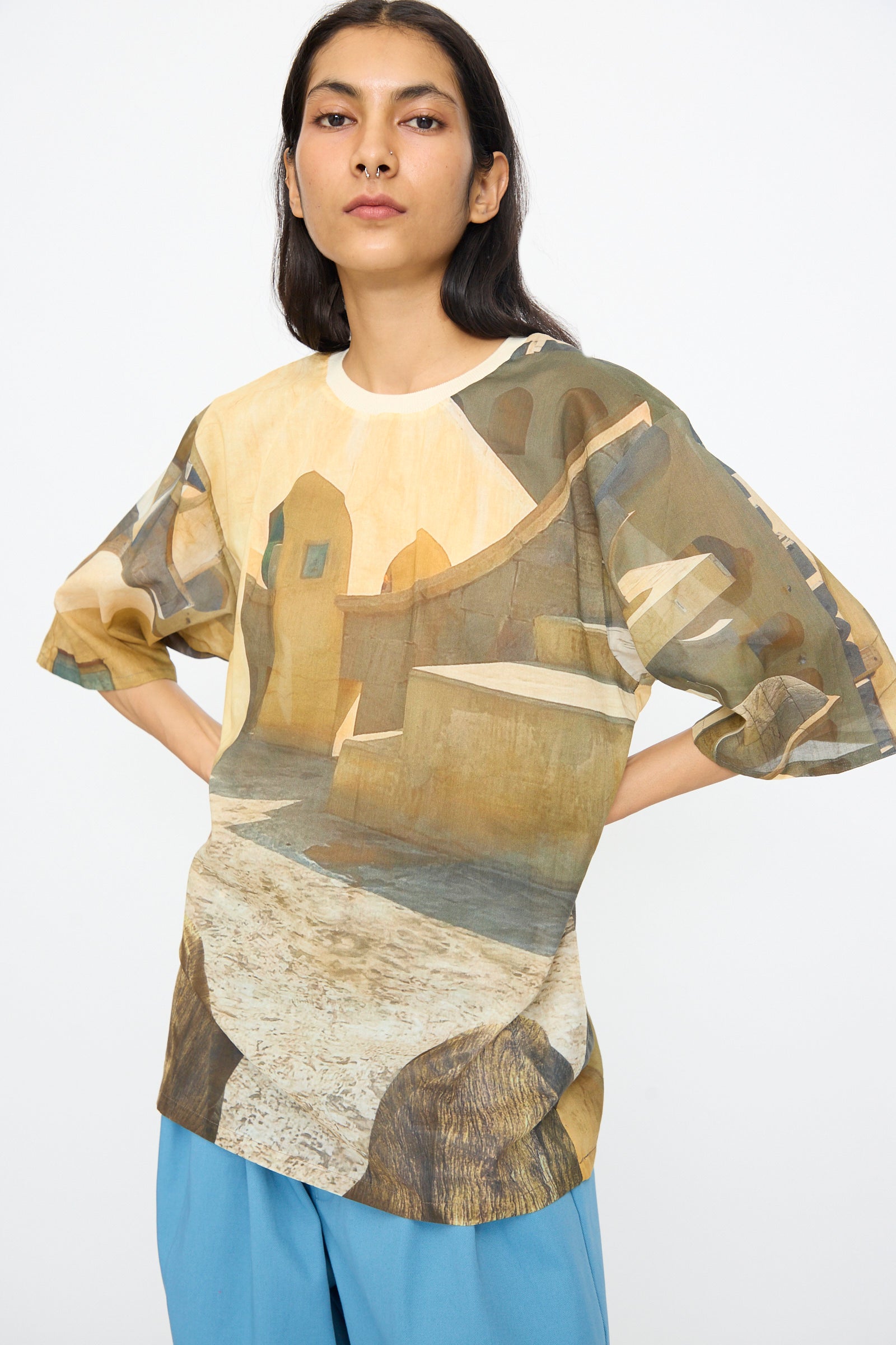 A person wearing the Bless Holidayjaipurobservatory Tee in Print, featuring an oversized boxy fit and architectural landscape design, with blue pants, stands hands on hips against a white background.