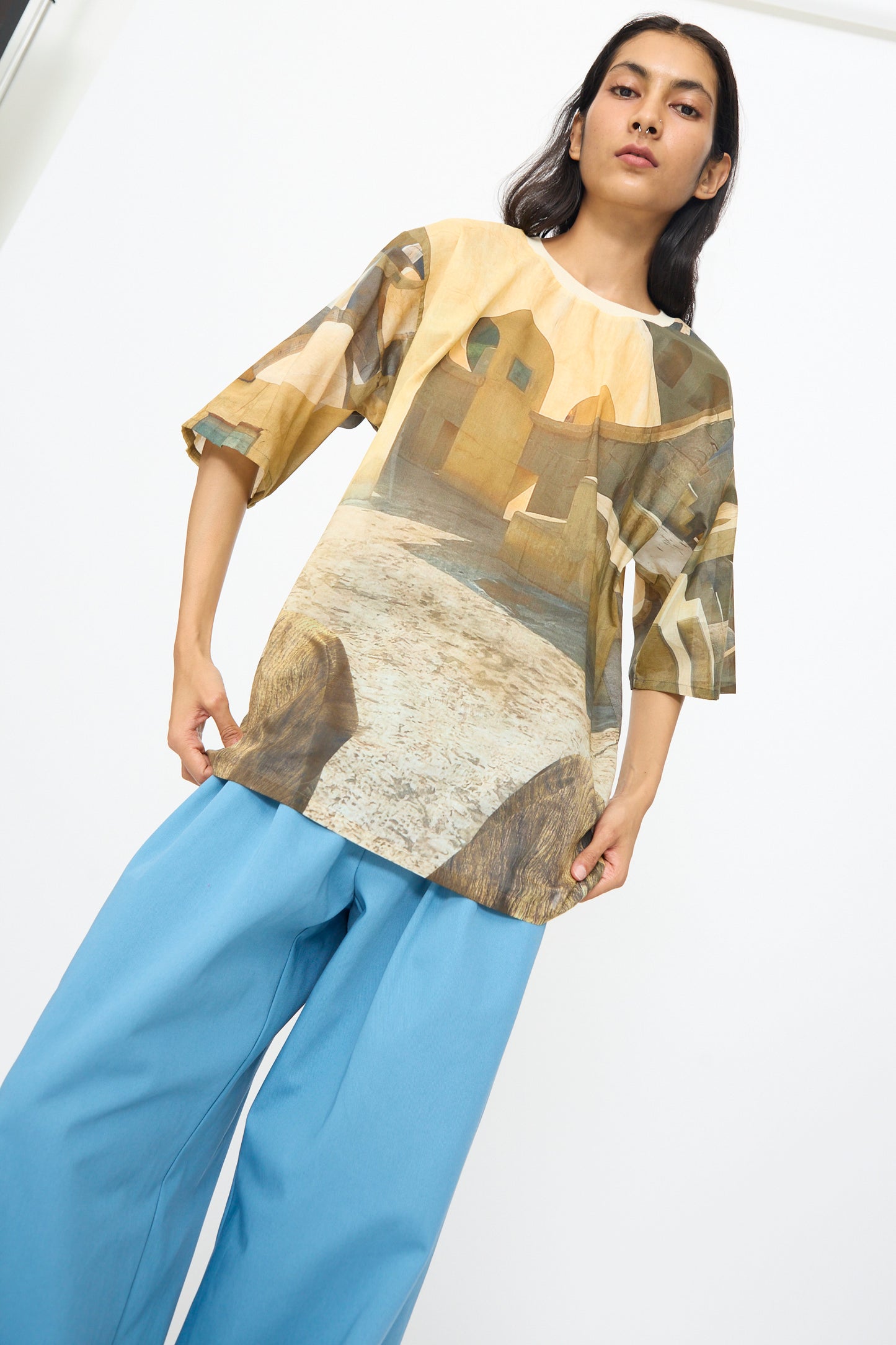 Indoors, a person wears the Bless Holidayjaipurobservatory Tee in Print—a semi-sheer cotton shirt with architectural patterns and oversized boxy fit—paired with loose blue pants.