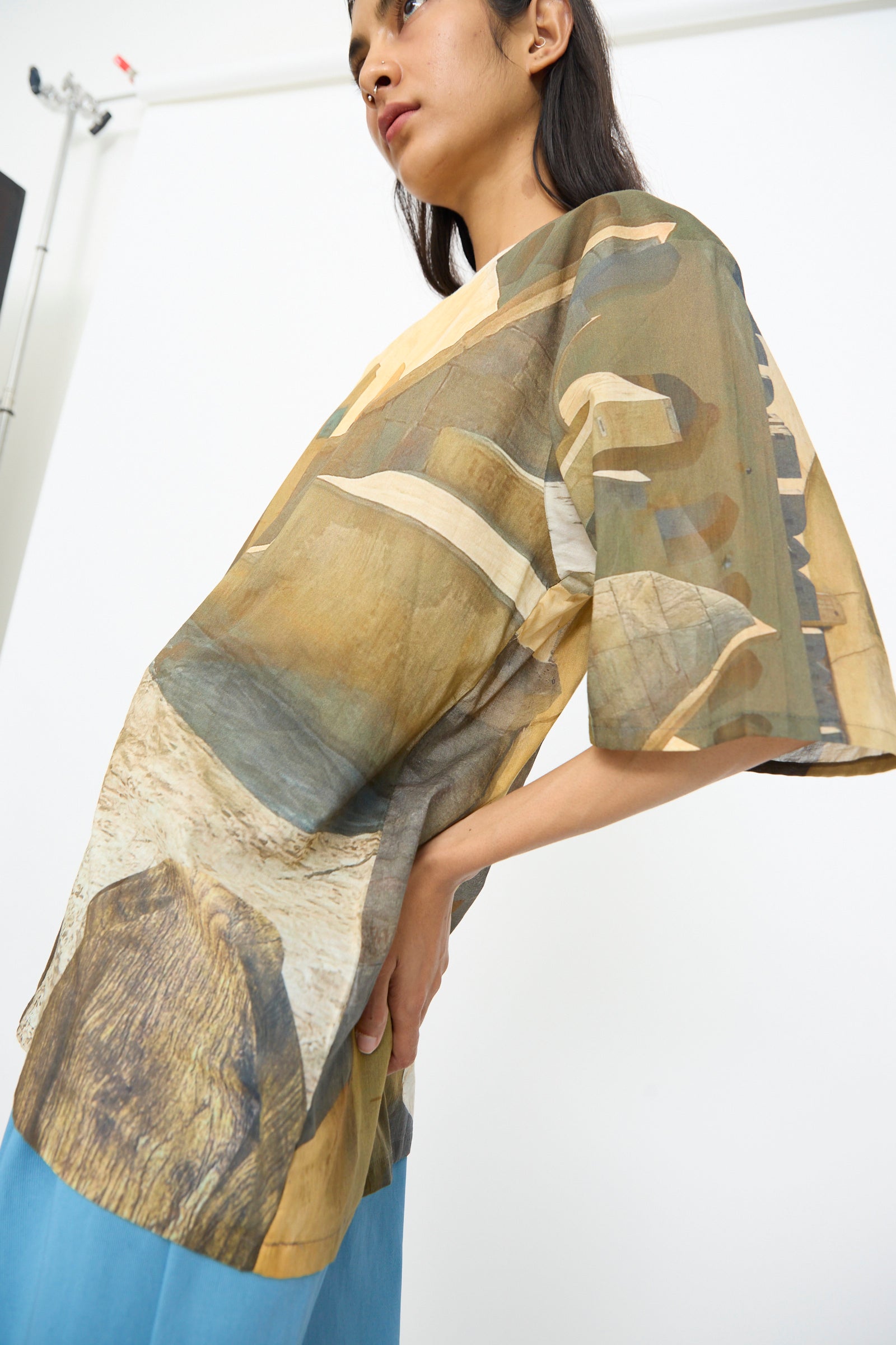 A person stands sideways in a Bless Holidayjaipurobservatory Tee in Print with an oversized boxy fit and abstract earth-tone pattern, paired with blue pants, set against a plain white background.