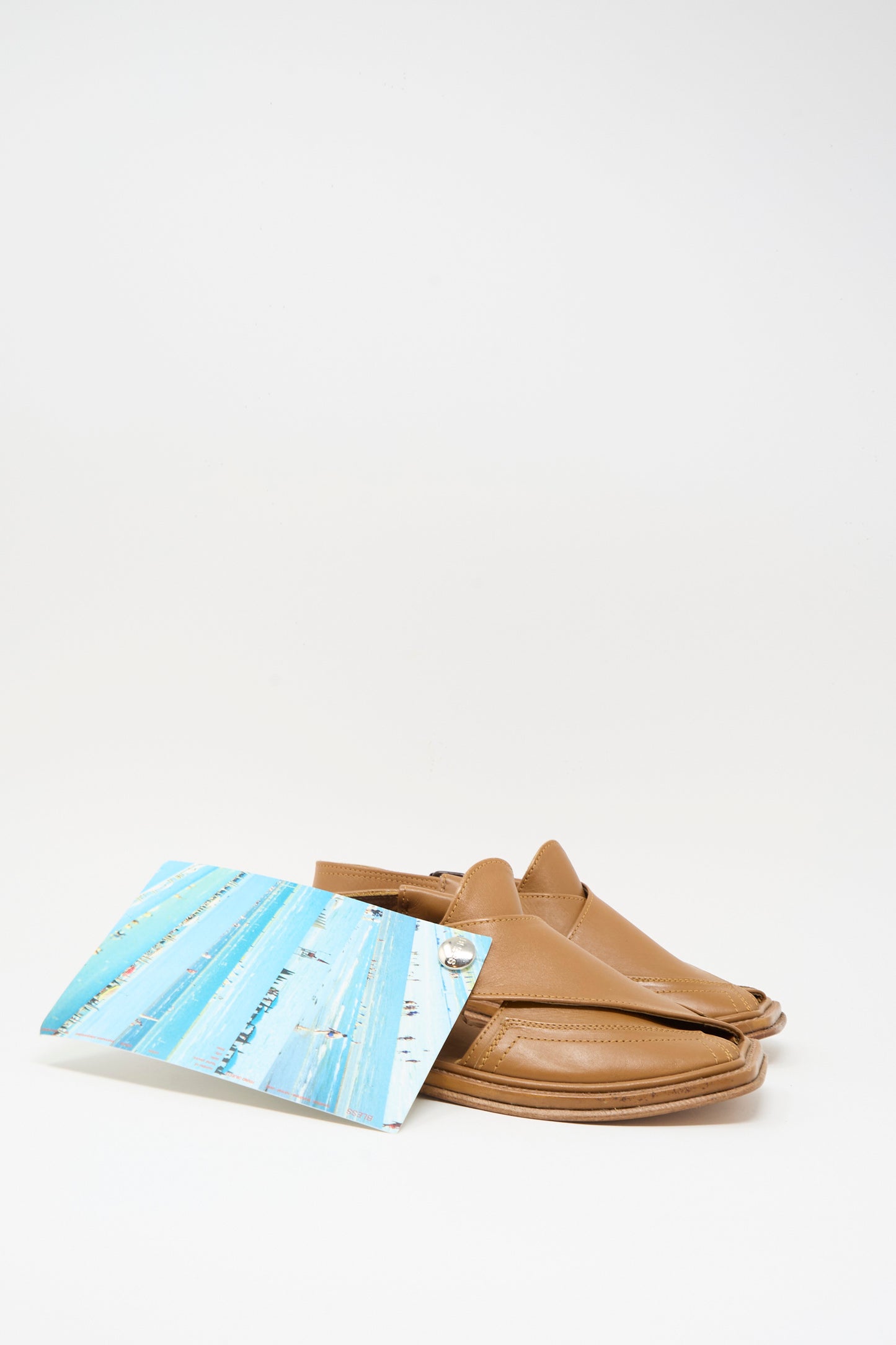 The Bless No. 08 Desertsandals in Light Brown, featuring a crossover design, are shown next to a rectangular card with blue and white stripes.