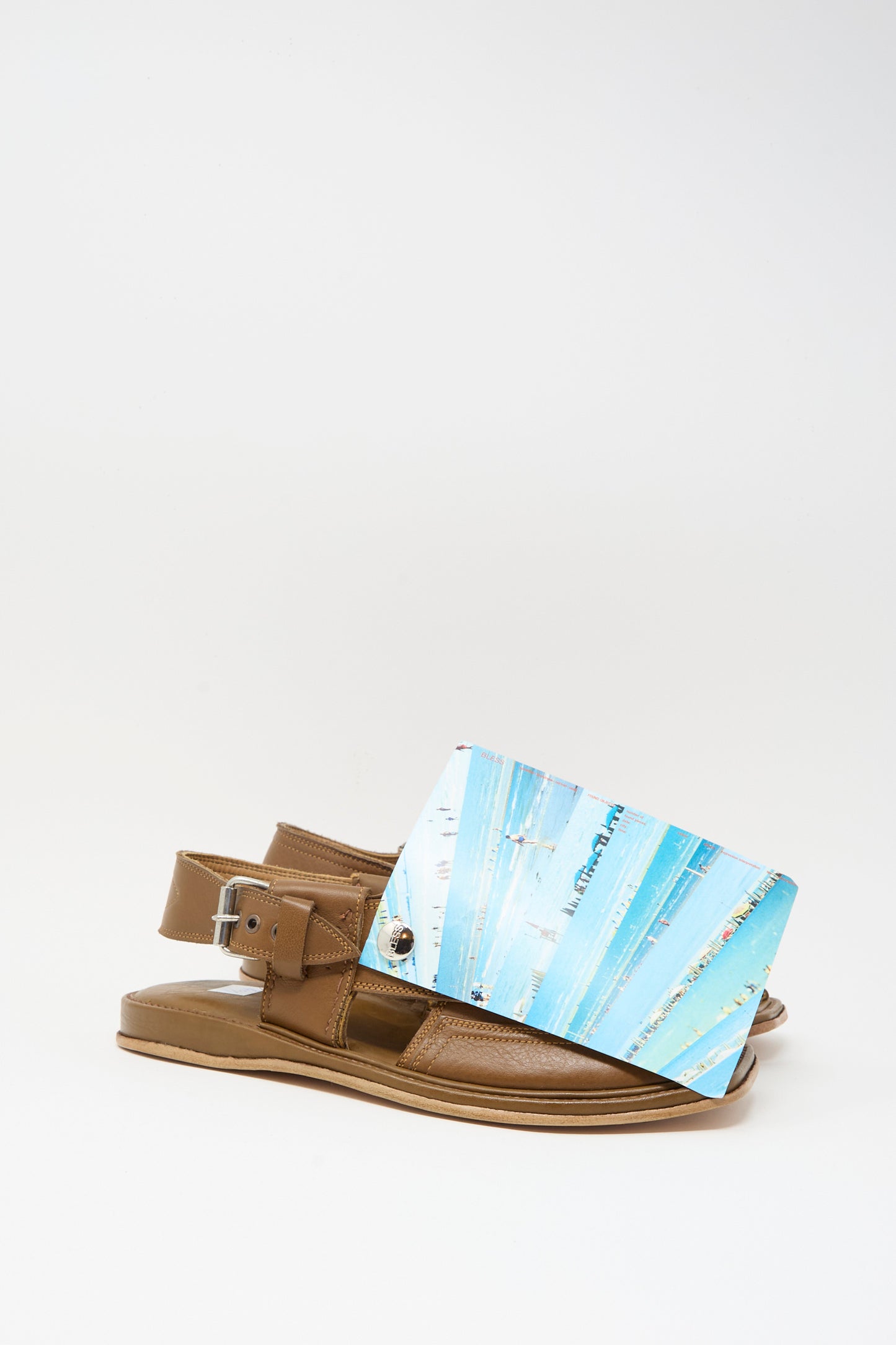 A pair of Bless No. 08 Desertsandals in dark brown, featuring an adjustable back strap, are partially covered by a blue and white rectangular card with decorative lines and text.