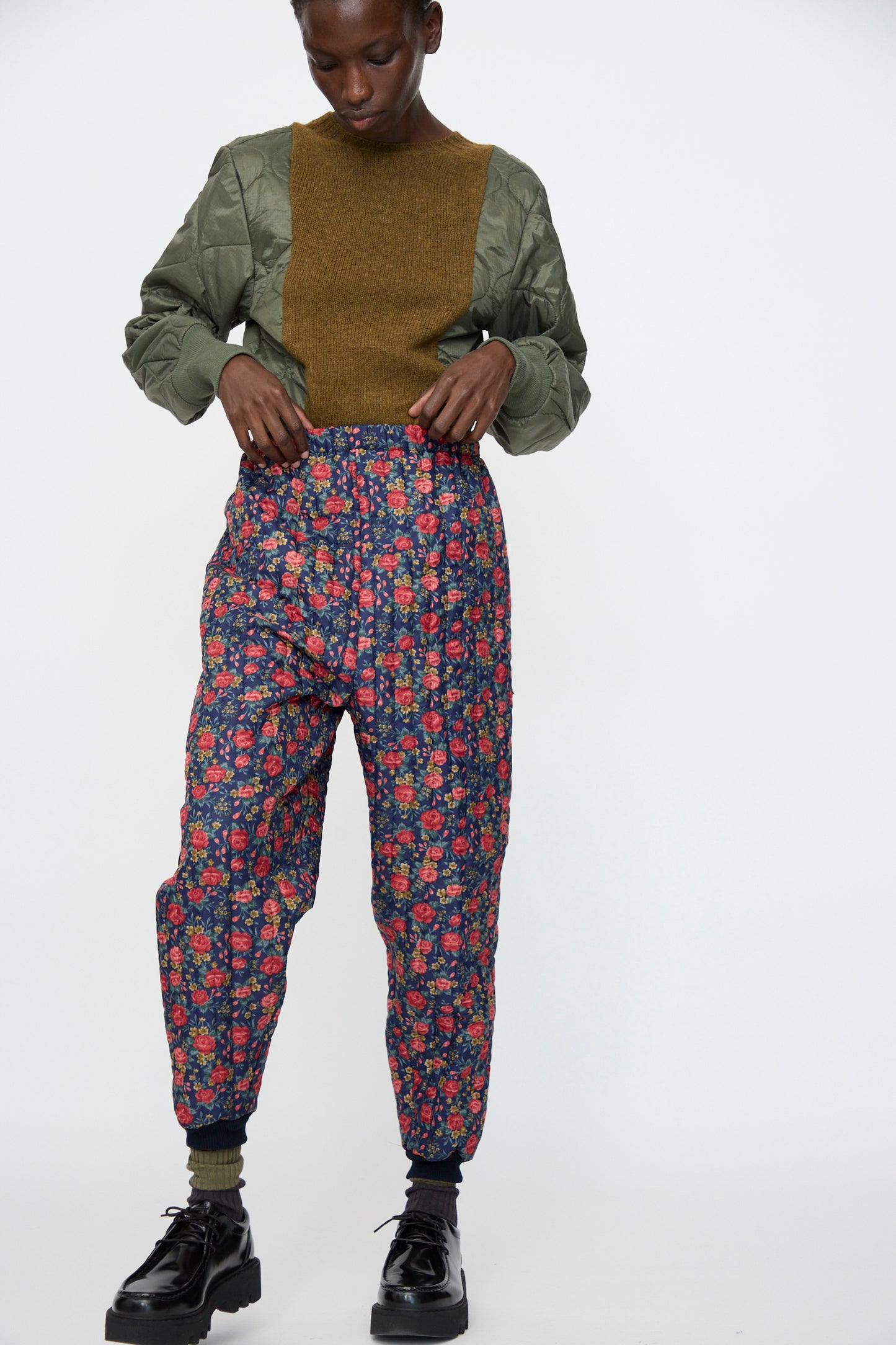A person wears the Bless No. 08 Monpe Pant in Floral Print I with a green quilted jacket and brown sweater, standing against a plain white background.