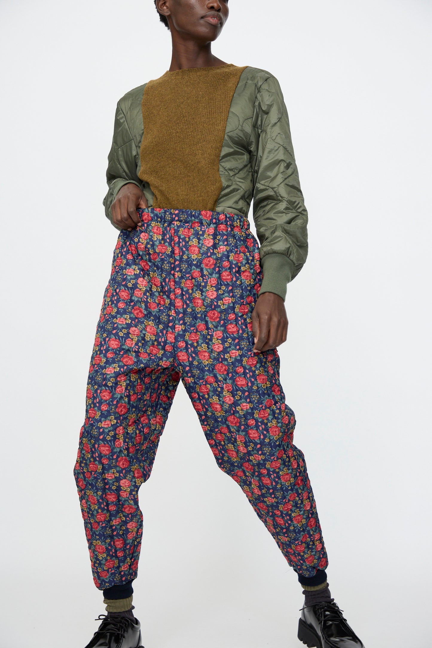 A person wears the Bless No. 08 Monpe Pant in Floral Print I with a long-sleeve olive green and brown top and black shoes, standing against a plain white background.