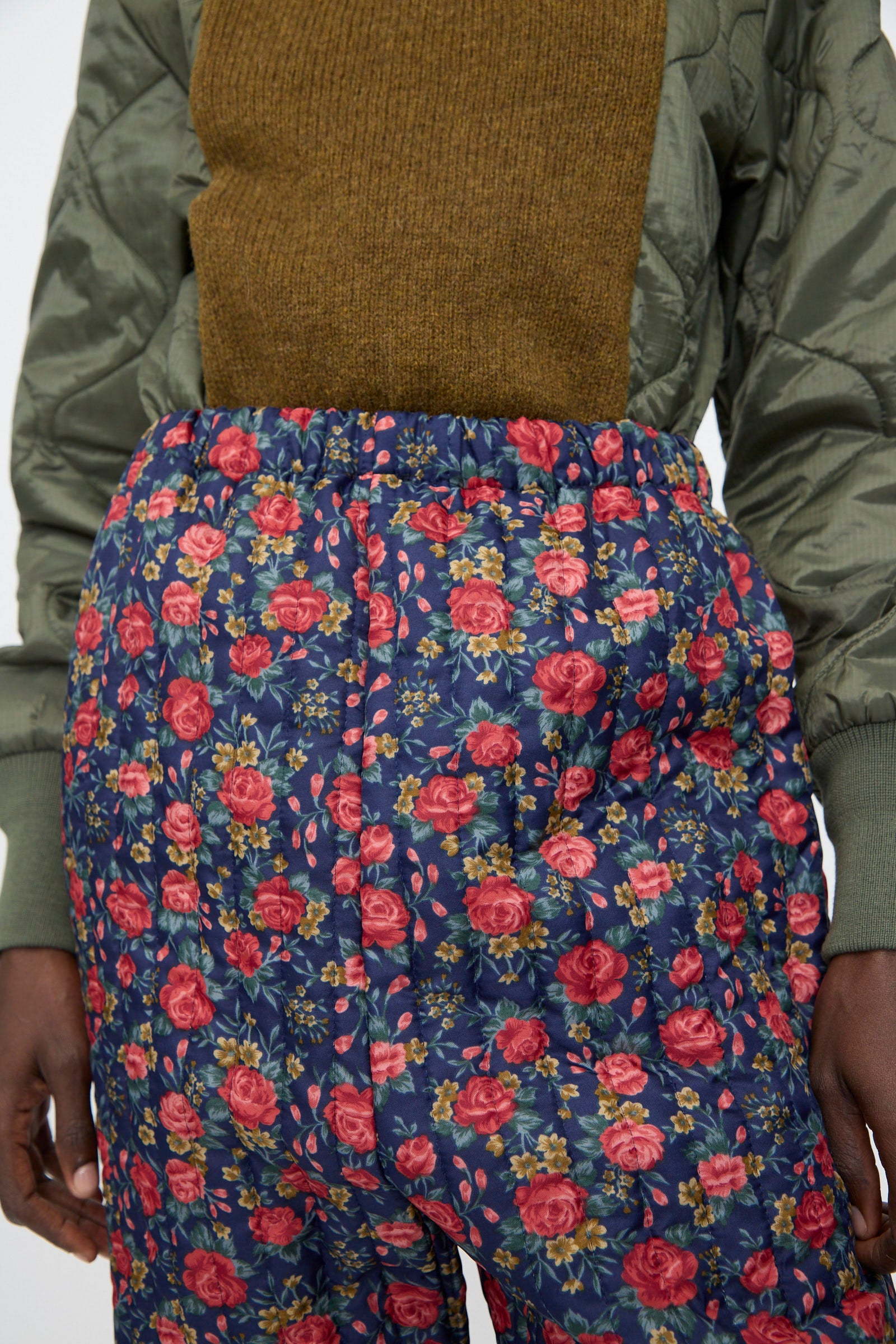 A person wearing a green quilted jacket, a brown sweater, and Bless No. 08 Monpe Pant in Floral Print I.