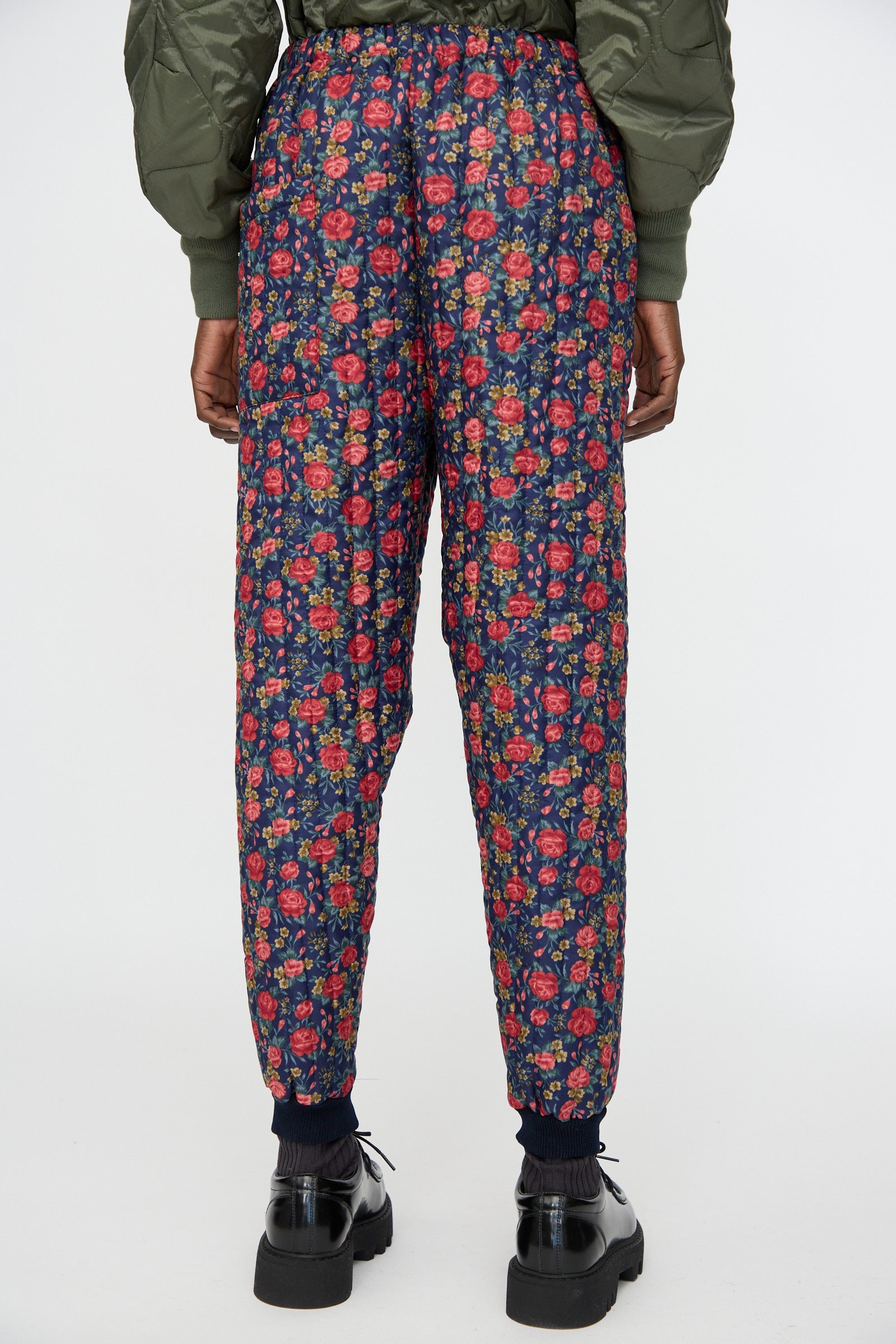 Person standing, seen from the back, wearing Bless No. 08 Monpe Pant in Floral Print I—navy blue workwear pants with a red floral pattern—paired with black shoes and a green quilted jacket.