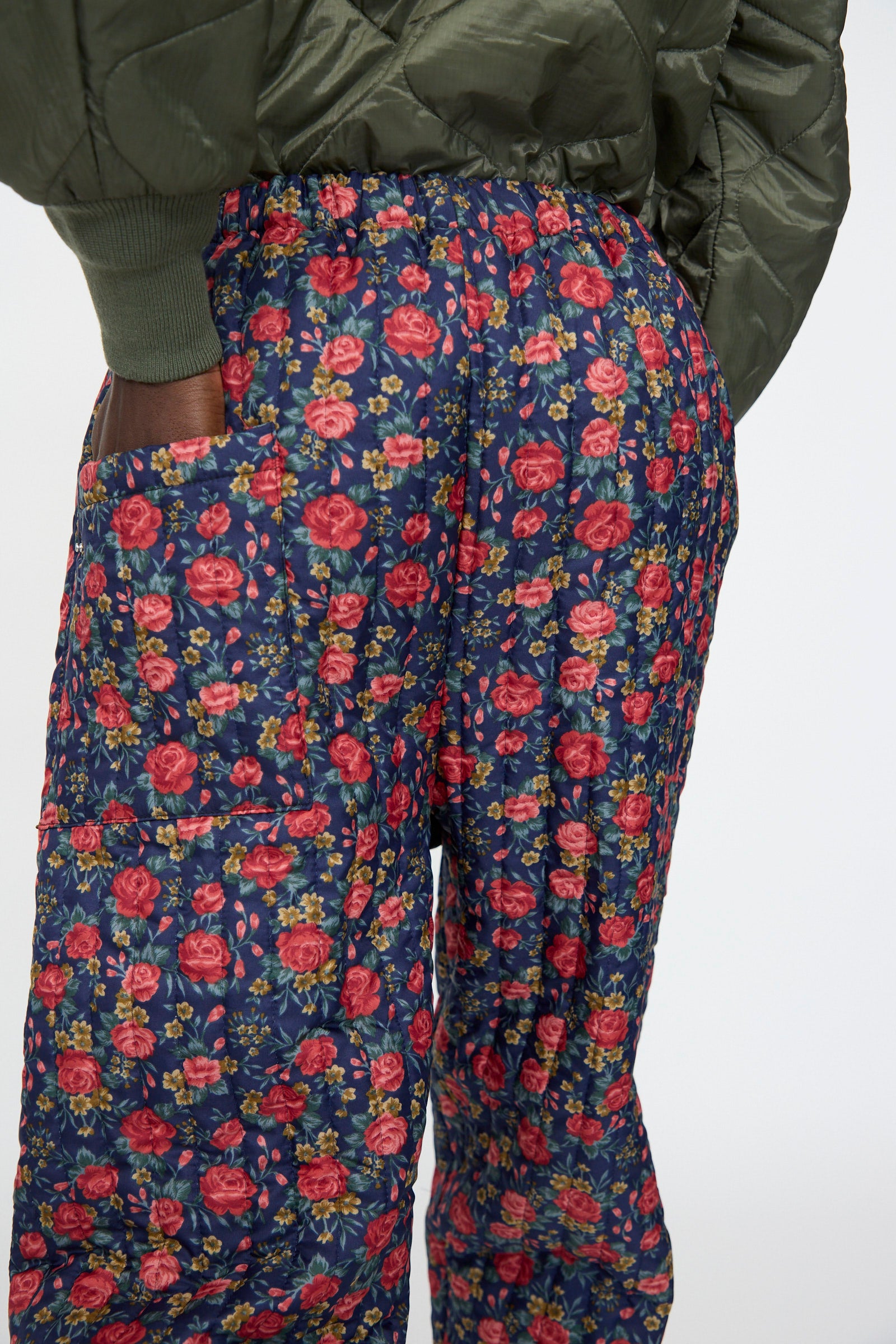 A person seen from the back with one hand in their pocket wears the Bless No. 08 Monpe Pant in Floral Print I—blue workwear pants inspired by Japanese monpe, featuring a red and pink pattern—and an olive green quilted jacket.