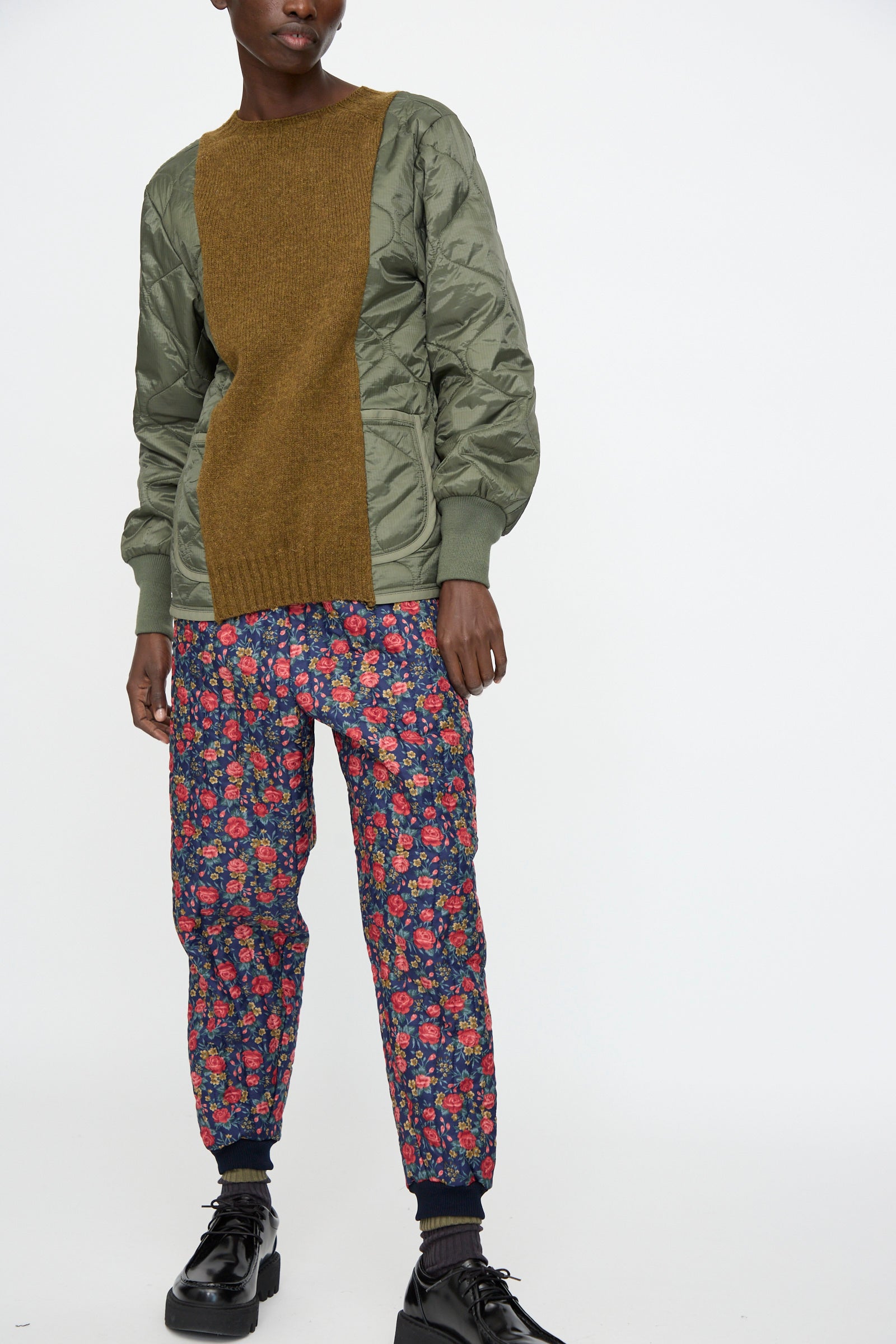 A person stands against a plain white background, wearing Bless’s No. 08 Monpe Pant in Floral Print I with an olive green and brown sweater with quilted sleeves and black shoes.