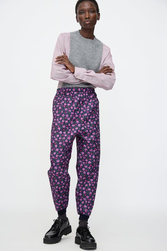 A person stands against a white background, wearing a gray and pink long-sleeve shirt, Bless No. 08 Monpe Pant in Floral Print II—purple floral Japanese nylon workwear pants—and black platform shoes.