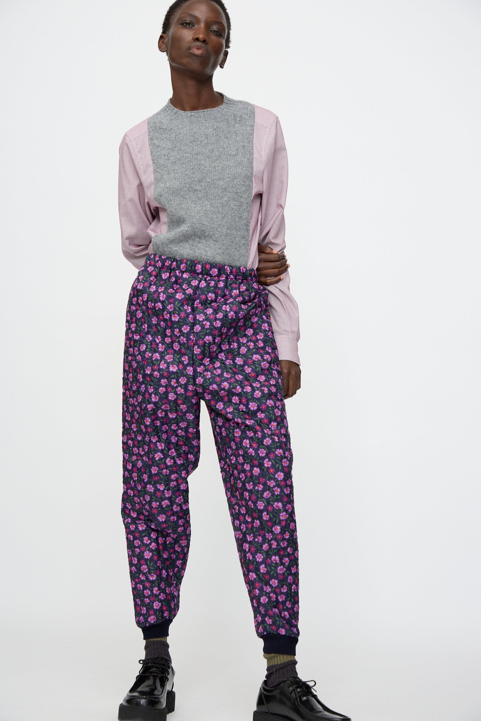 A person stands against a plain background wearing a grey top with pink sleeves, Bless No. 08 Monpe Pant in Floral Print II, and black shoes.