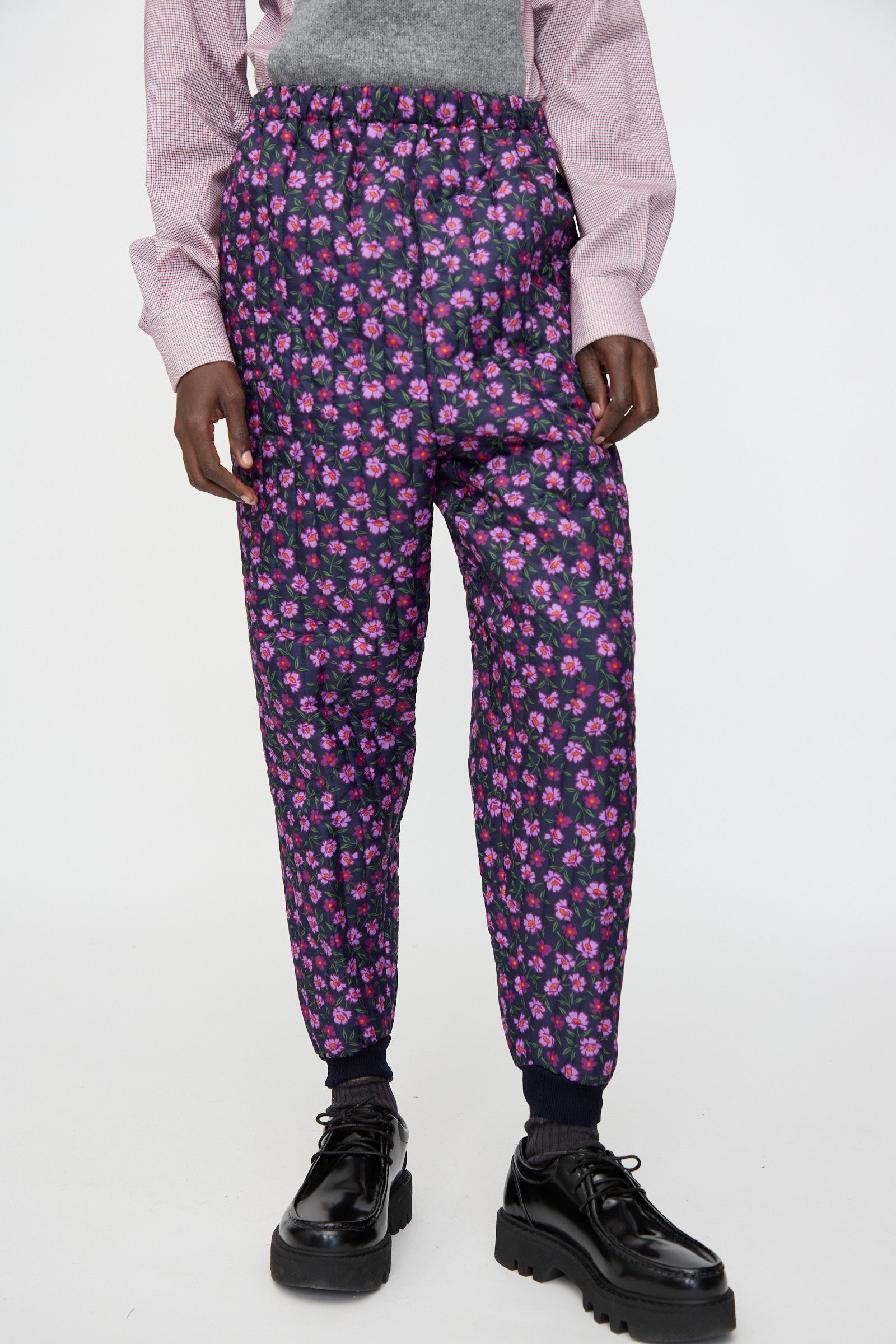 A person wears the Bless No. 08 Monpe Pant in Floral Print II—high-waisted, dark pants with a pink and purple floral pattern—paired with a light pink shirt and black platform shoes, standing against a white background.