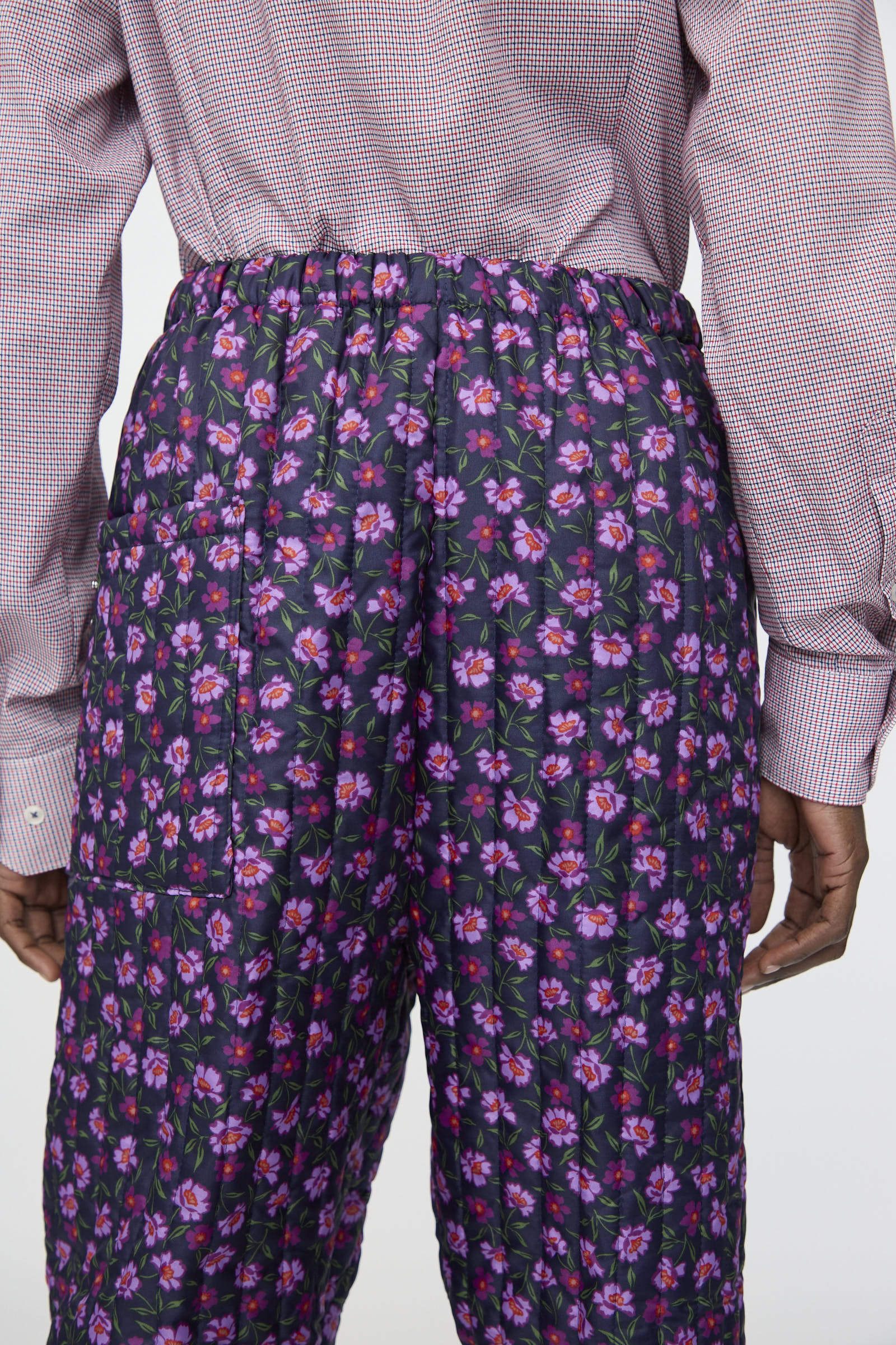 A person seen from the back wears a light red checkered shirt and Bless’s No. 08 Monpe Pant in Floral Print II, featuring a high-waisted fit with a pink and purple floral pattern.