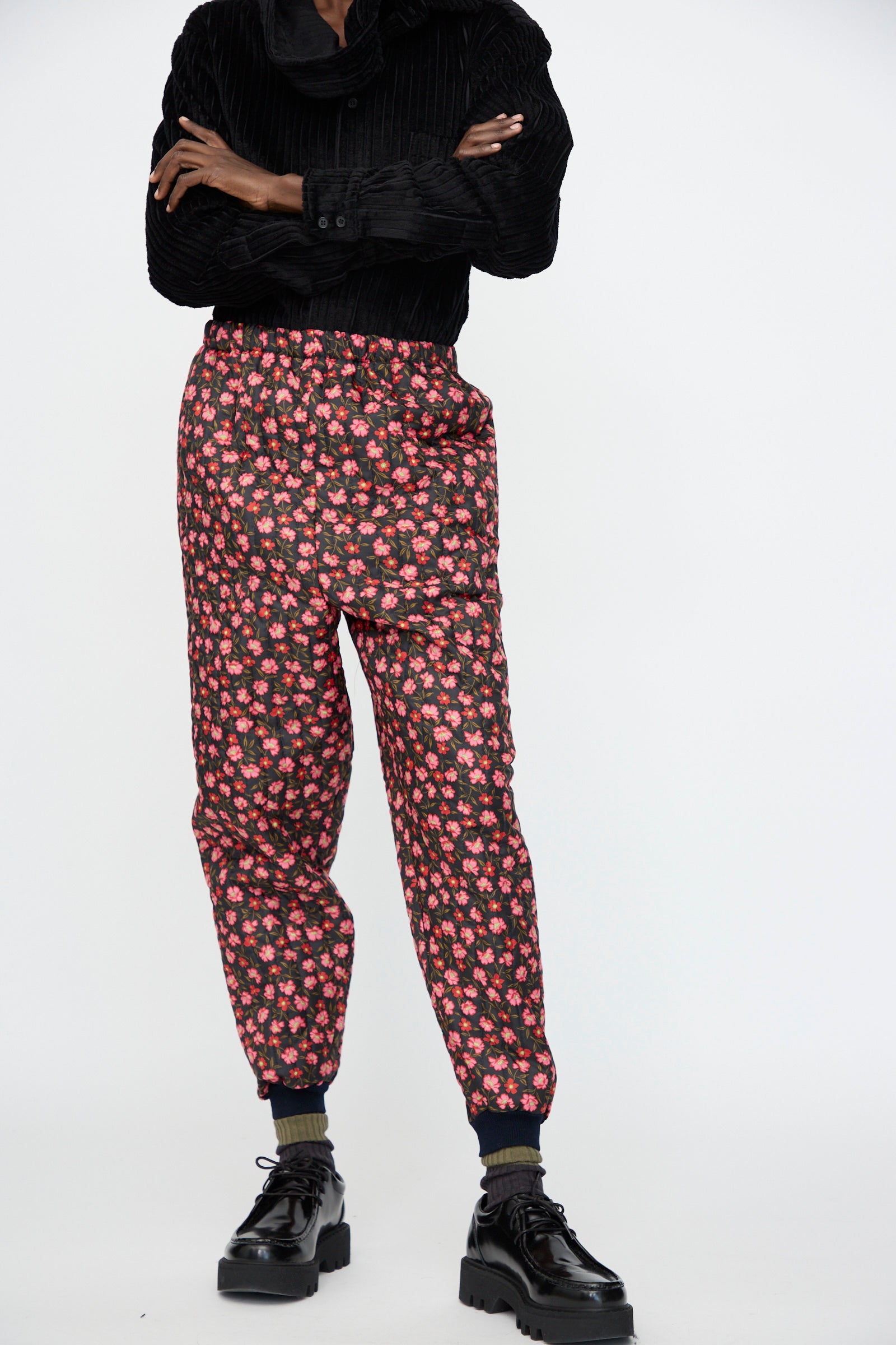 A person stands with arms crossed against a plain background, wearing a black textured shirt, Bless No. 08 Monpe Pant in Floral Print IV, dark socks, and black platform shoes.