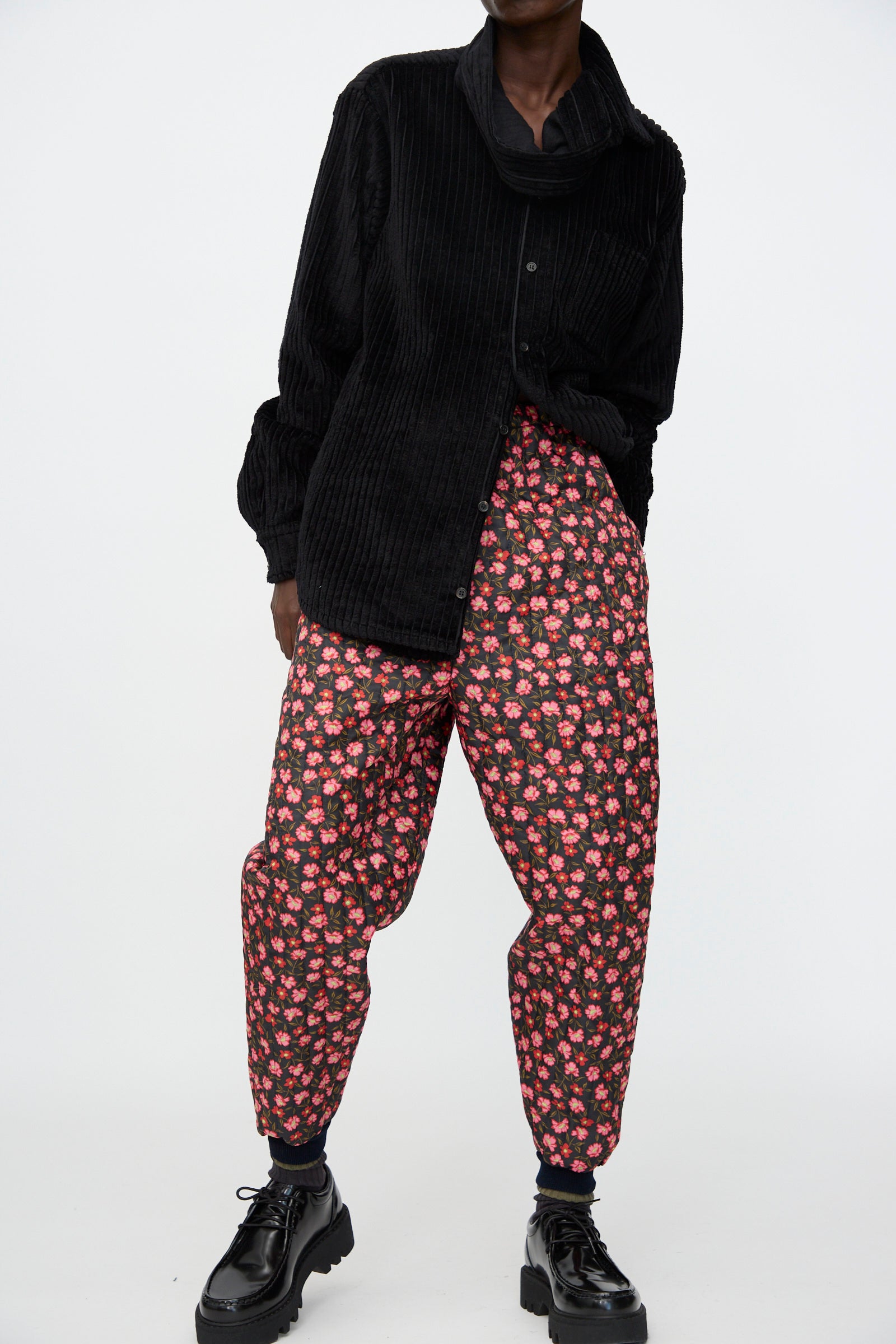A person stands against a plain white background wearing a loose black corduroy shirt, black socks, black patent leather shoes, and the Bless No. 08 Monpe Pant in Floral Print IV.