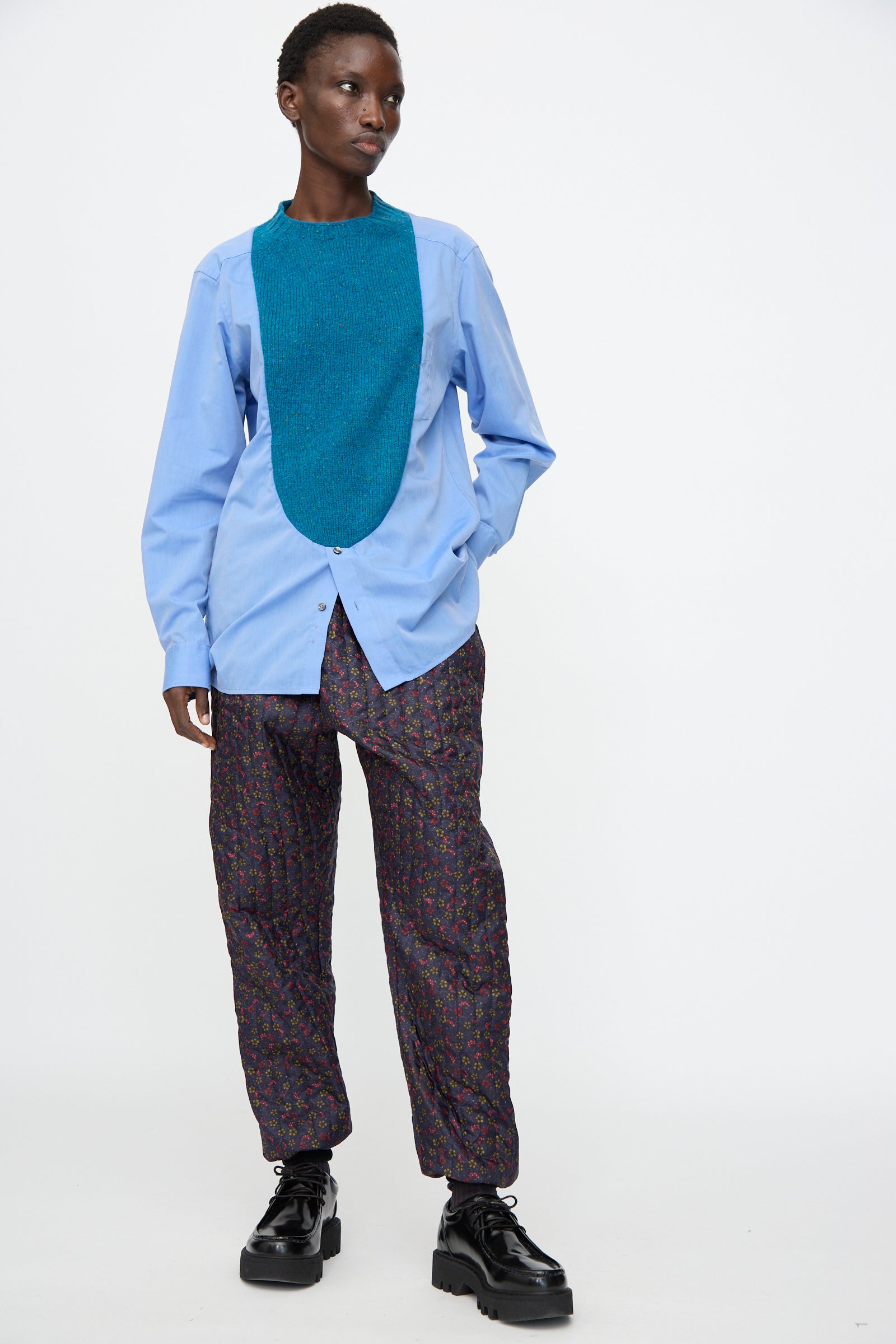 A person wears a light blue shirt, teal bib, Bless No. 08 Monpe Pant in Floral Print V inspired by Japanese workwear, and black platform shoes, standing against a plain white background.