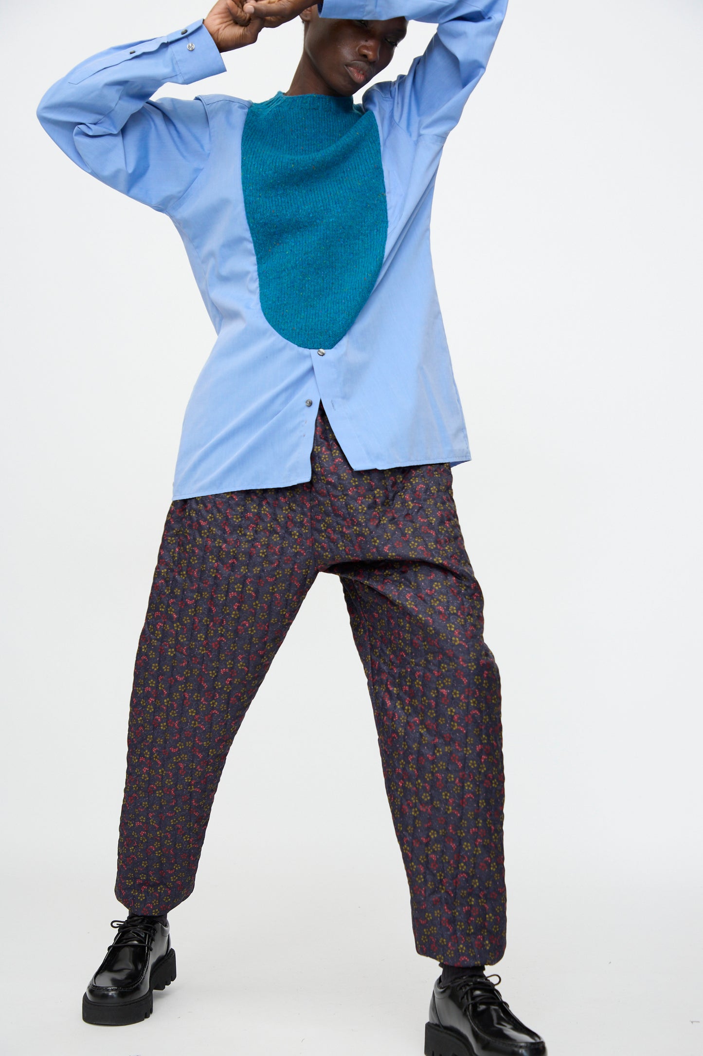 Person posing in a light blue shirt, teal bib, Bless No. 08 Monpe Pant in Floral Print V, black platform shoes, and a blue hat against a plain white background.