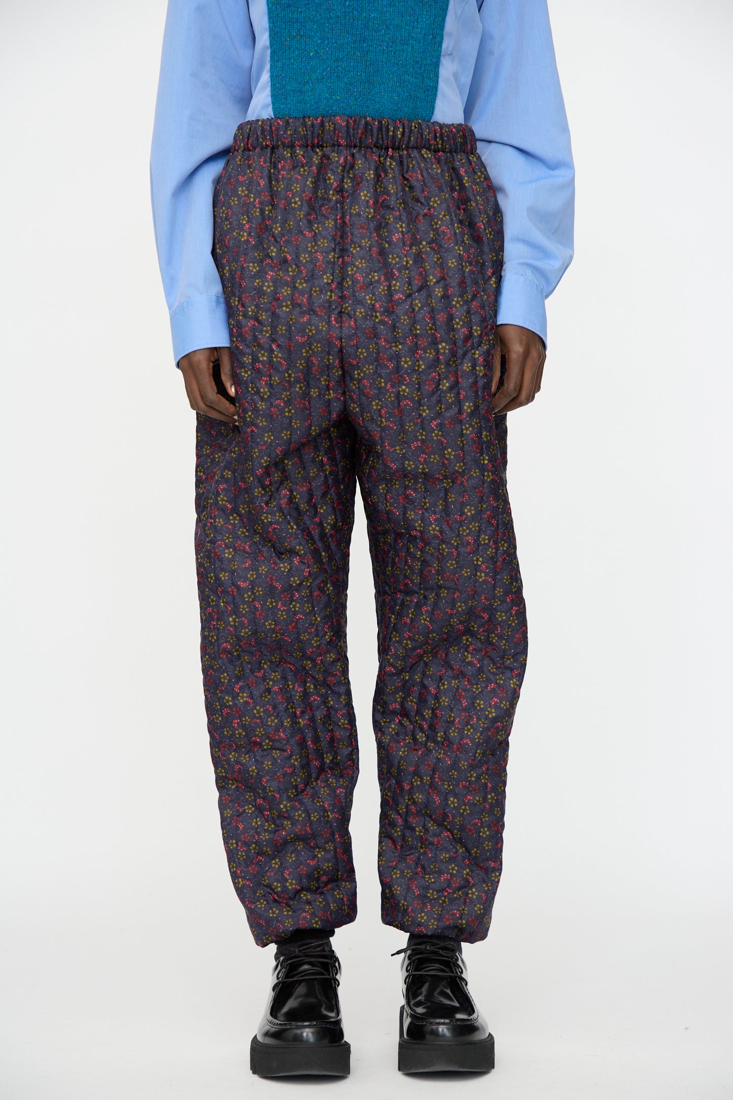 A person wearing a light blue shirt, teal vest, Bless No. 08 Monpe Pant in Floral Print V, and black platform shoes.