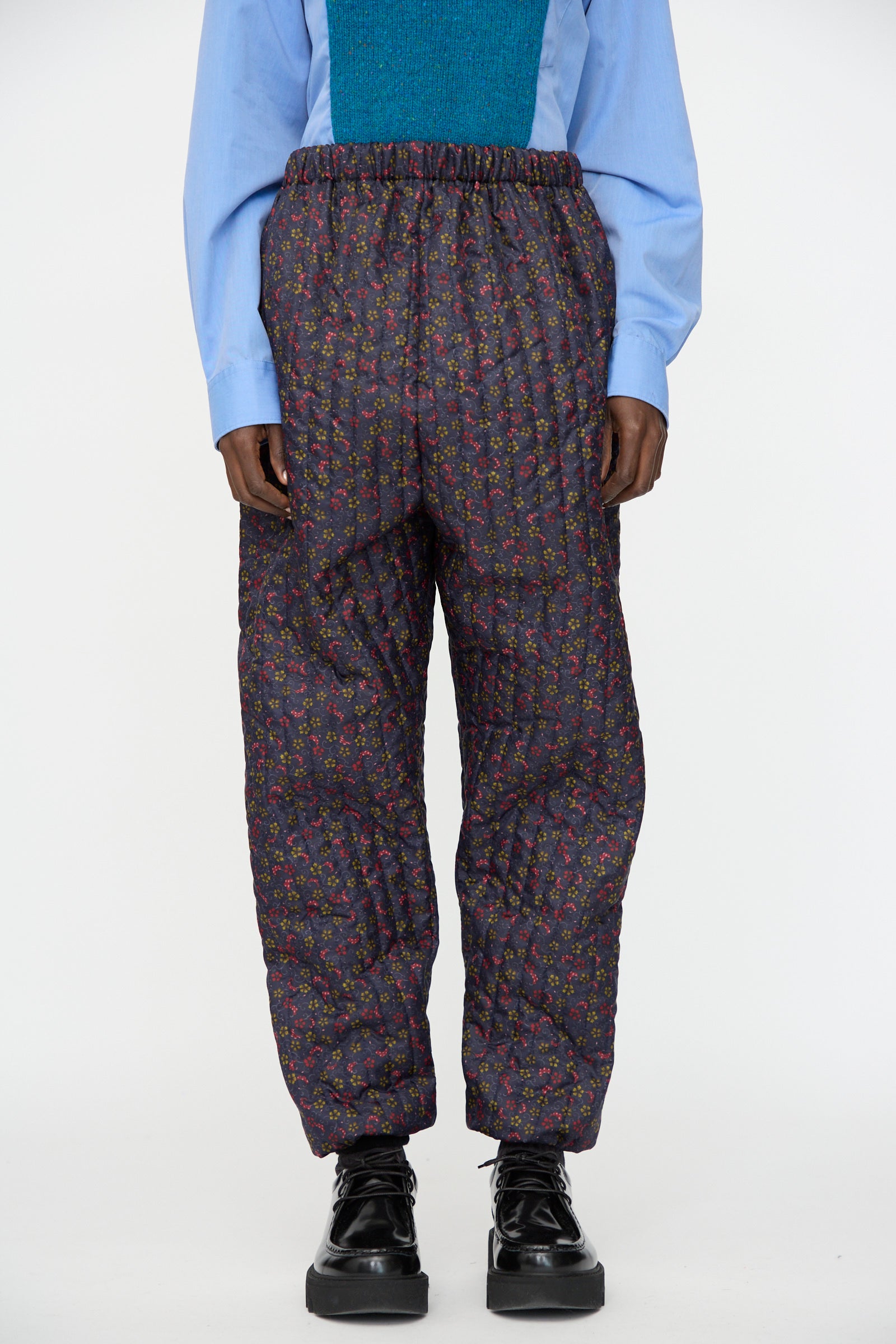 A person wearing a light blue shirt, teal vest, Bless No. 08 Monpe Pant in Floral Print V, and black platform shoes.