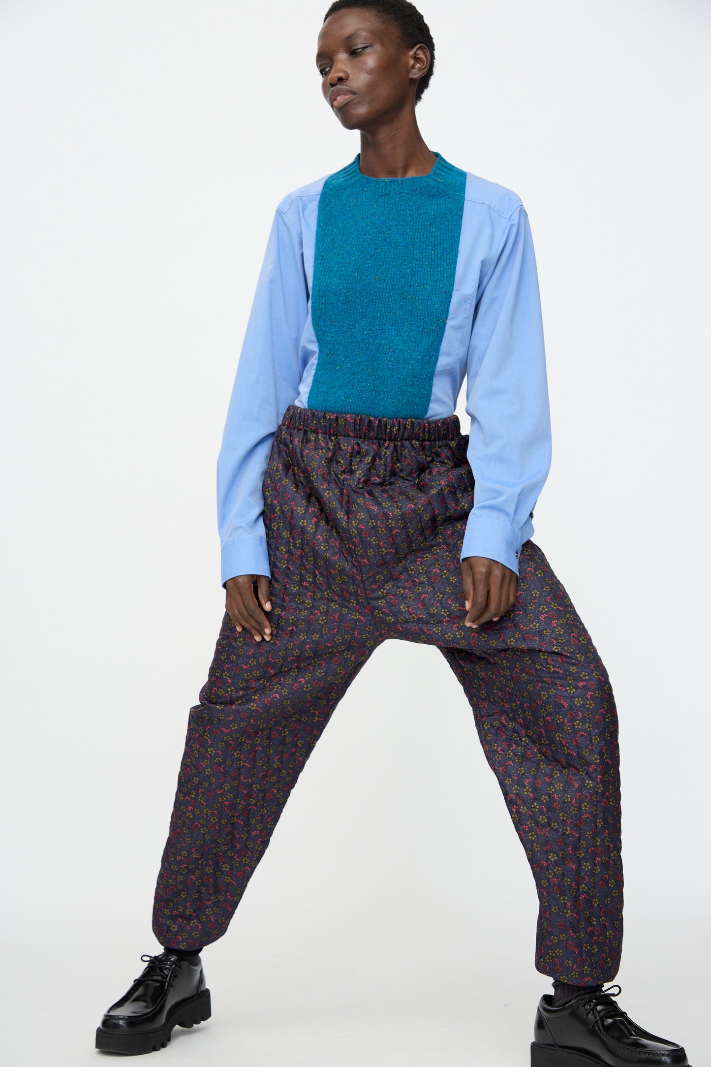 A person stands wide-legged against a plain white background, wearing Bless’s No. 08 Monpe Pant in Floral Print V with black shoes and a blue long-sleeve top featuring a teal panel.