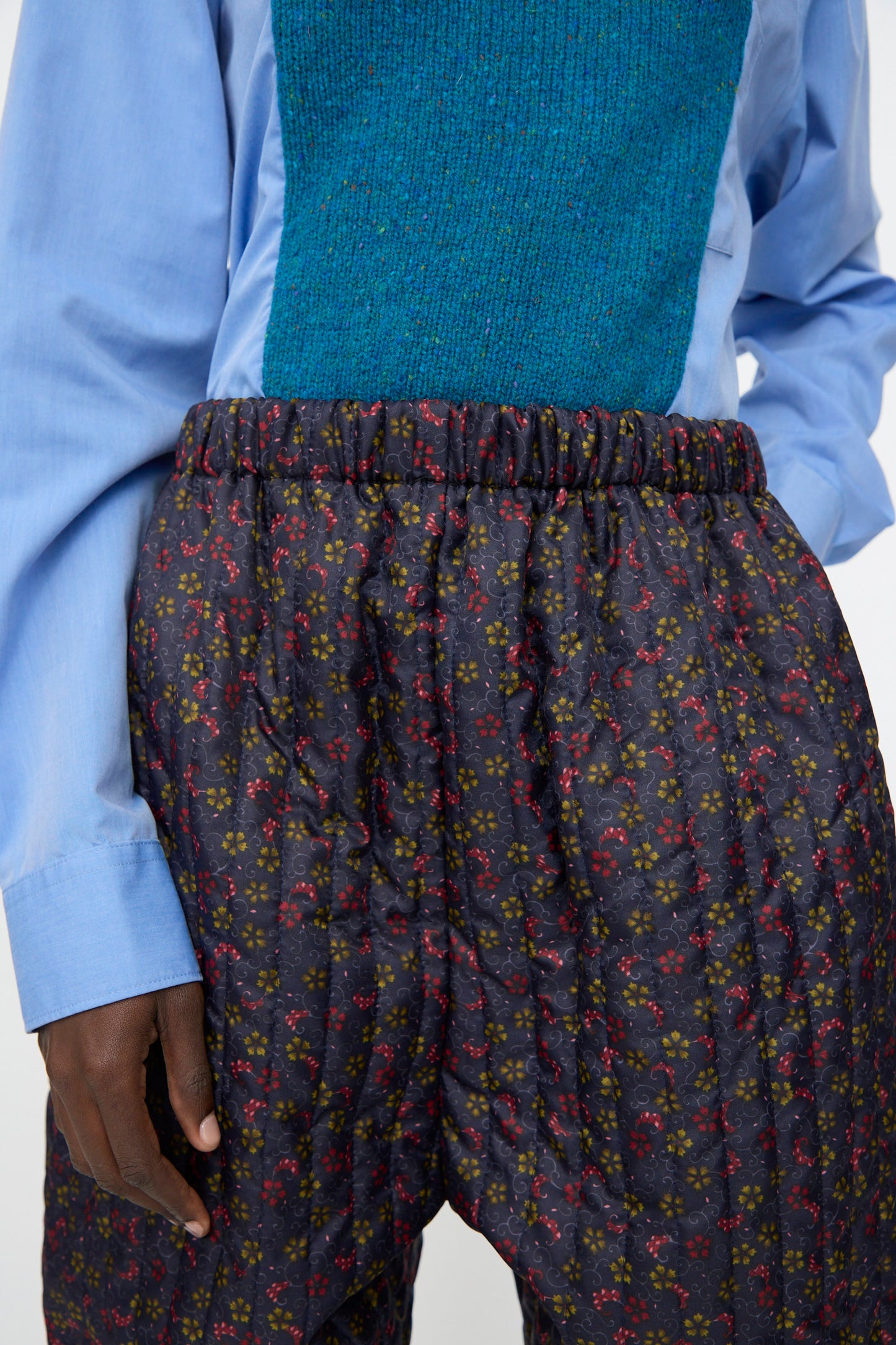 A person wears Bless’s No. 08 Monpe Pant in Floral Print V—navy, high-waisted, floral nylon pants inspired by Japanese workwear—paired with a blue shirt and teal textured vest.