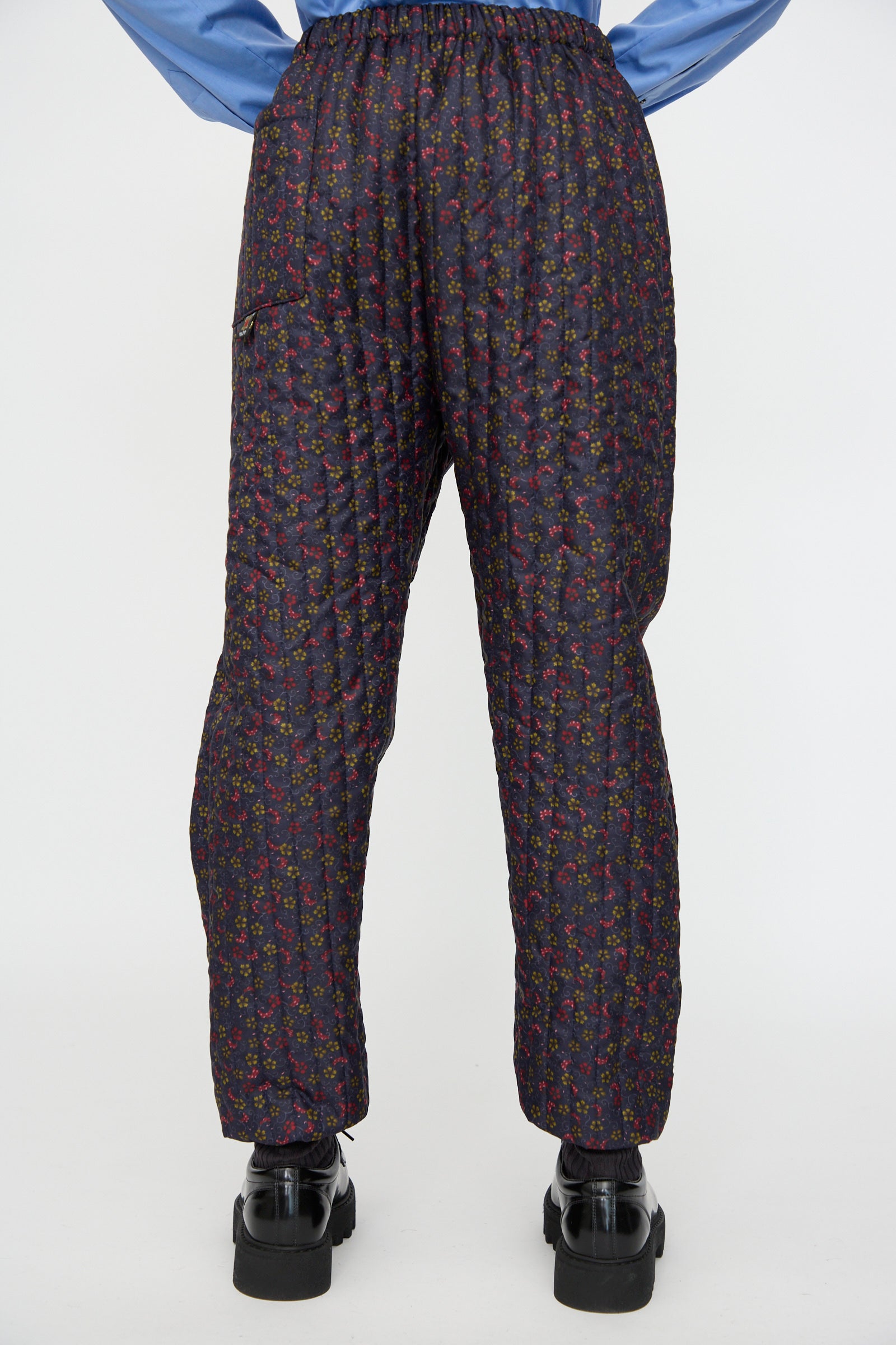 Person wearing the Bless No. 08 Monpe Pant in Floral Print V, paired with black shoes and a light blue shirt, photographed from the back against a white background.