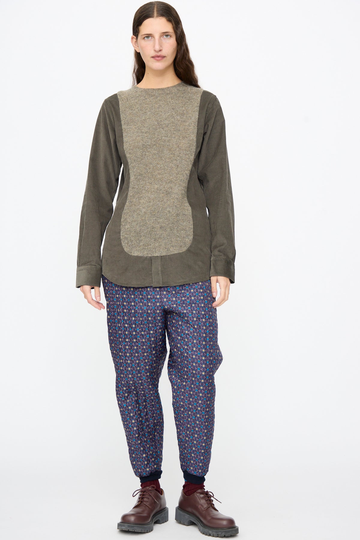 A person wearing a long-sleeved olive shirt with a beige panel, Bless No. 08 Monpe Pants in Floral Print II, and brown lace-up shoes.