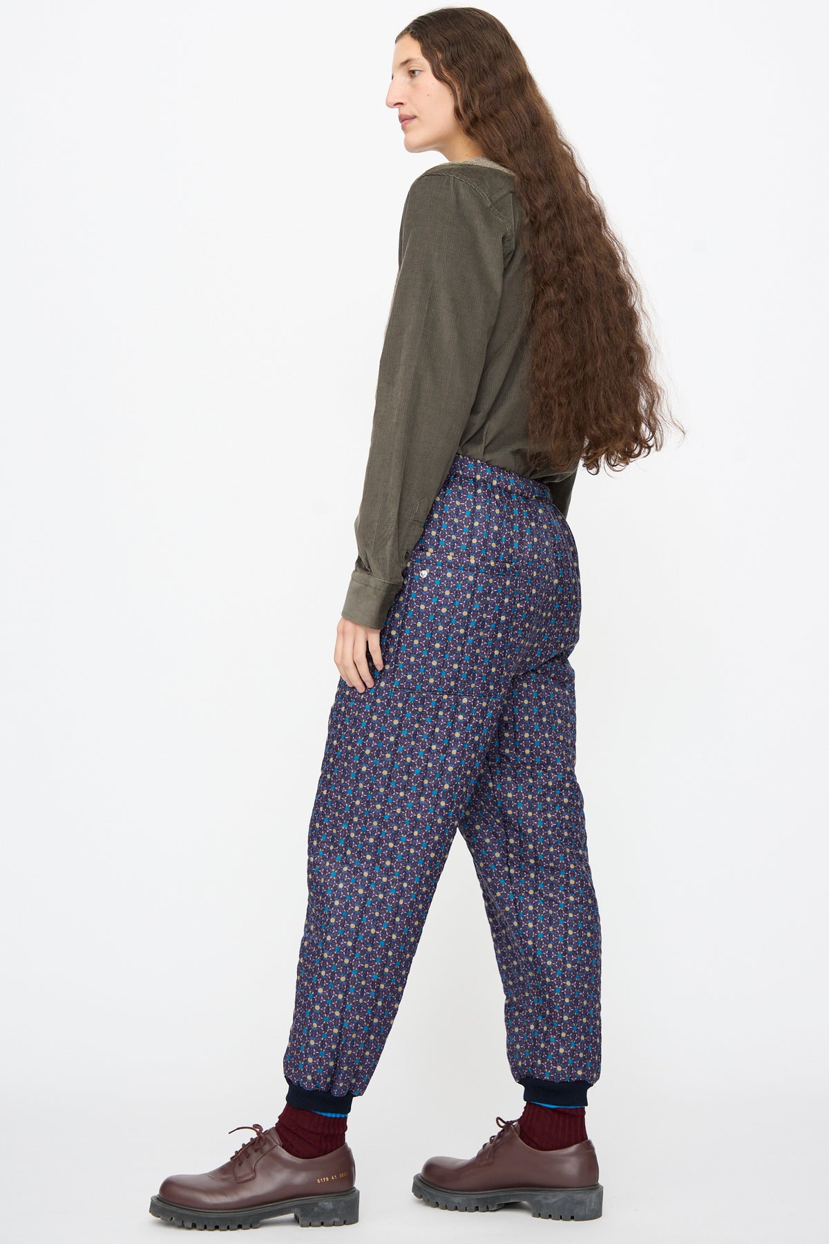A person wears a dark olive long-sleeved shirt, Bless No. 08 Monpe Pants in Floral Print II with ribbed cuffs, and brown leather lace-up shoes.